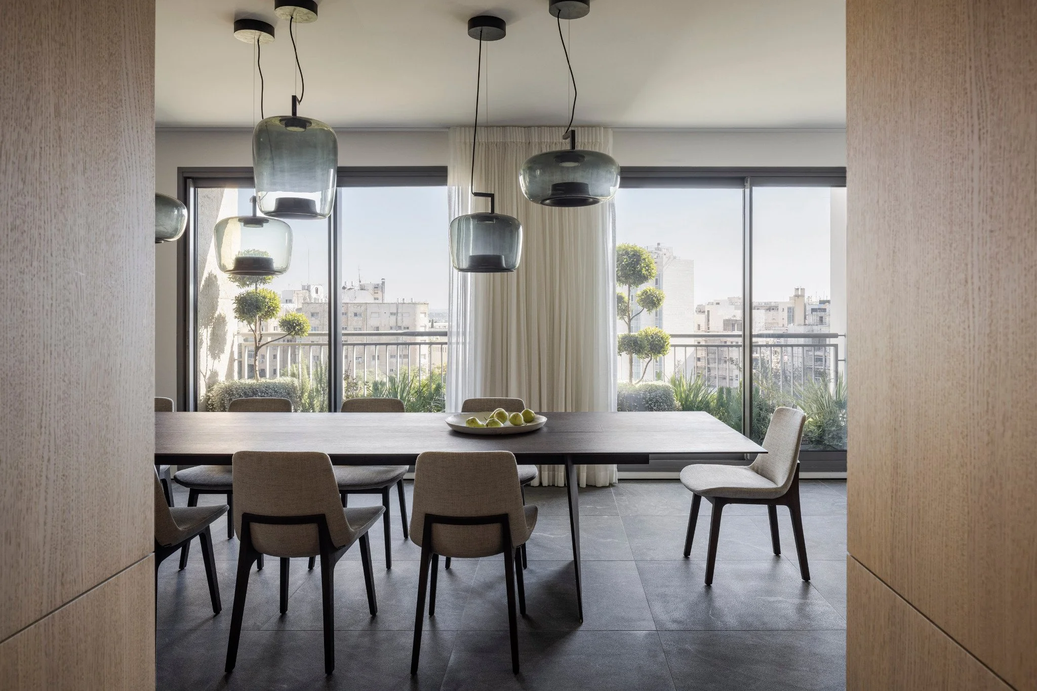 Modern dining room with a large rectangular table, eight beige upholstered chairs, and a white plate with green apples. Floor-to-ceiling windows with white curtains offer a city view and outdoor greenery. Black pendant lights hang above the table.