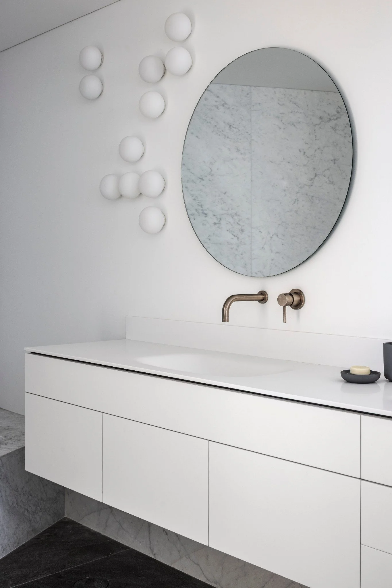 A modern bathroom with a white floating vanity, a round mirror with a thin black frame, a marble backsplash, a brushed nickel faucet, and a soap dish.