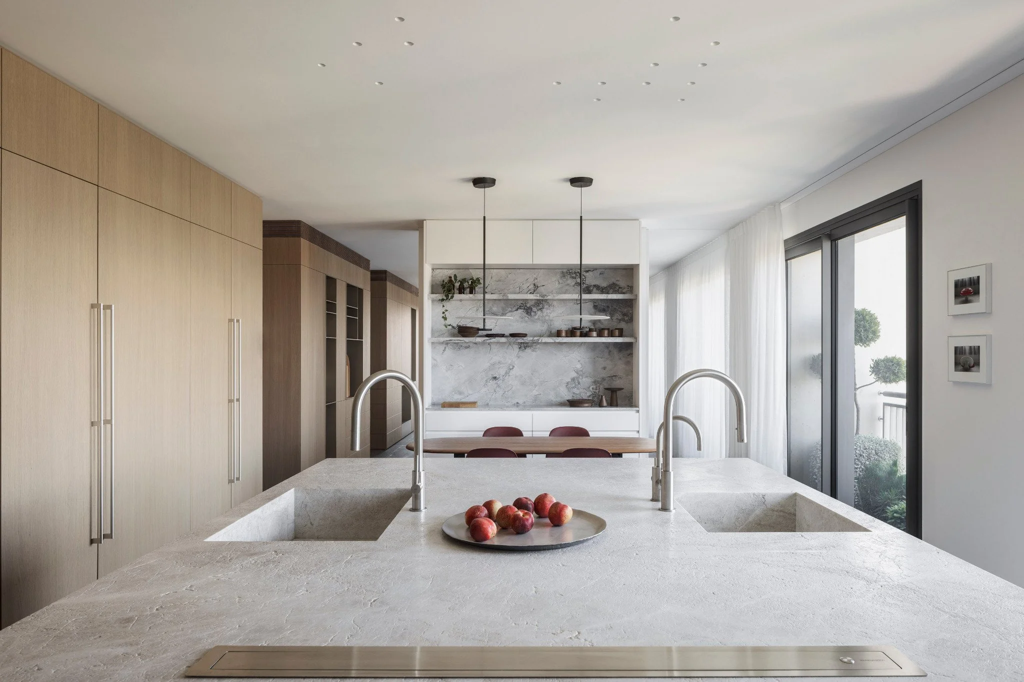 A modern kitchen with a large island in the foreground, apples in a bowl, beige wooden cabinets to the left, a marble backsplash with shelves and decorative items in the background, and large windows with white curtains to the right.