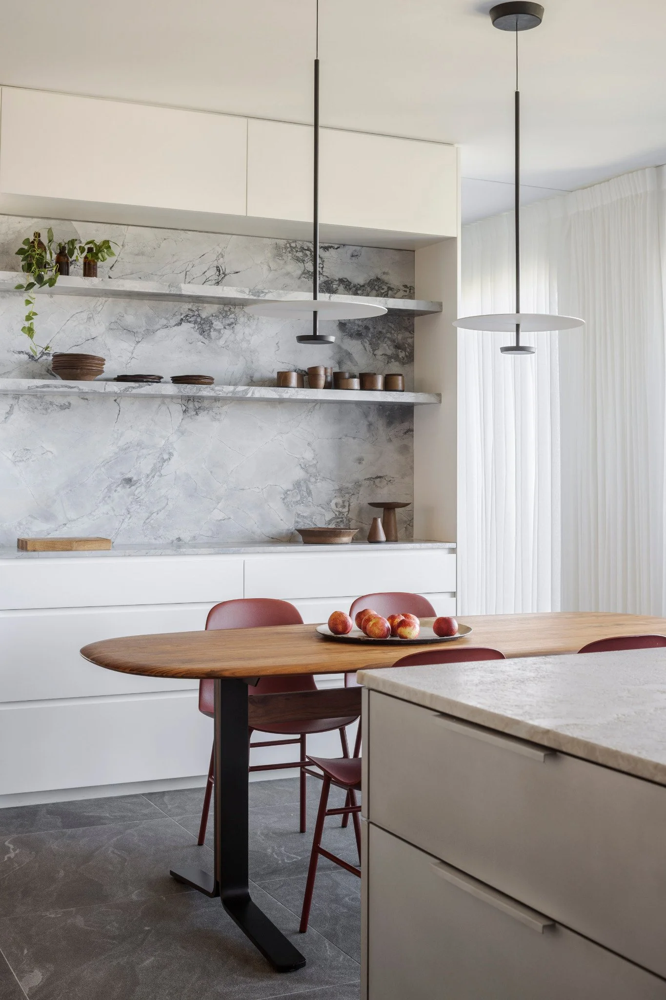 A modern kitchen with a wooden dining table, red chairs, a countertop with apples, marble backsplashes, white cabinetry, and contemporary pendant lights.