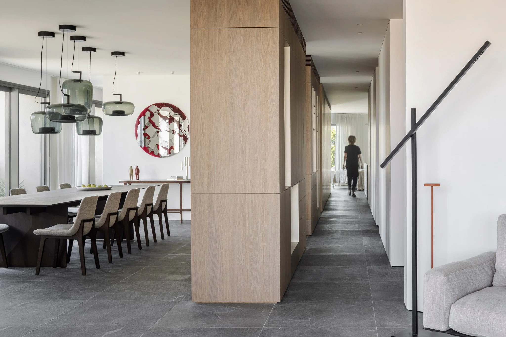 Modern dining and living area with hanging light fixtures, a circular mirror, wooden wall, hallway, and a person walking in the background.