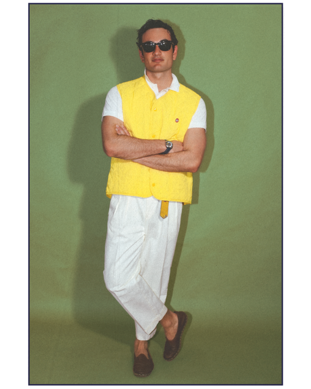 A man wearing sunglasses, a yellow vest over a white shirt, white pants, and dark shoes, standing against a green background with arms crossed.