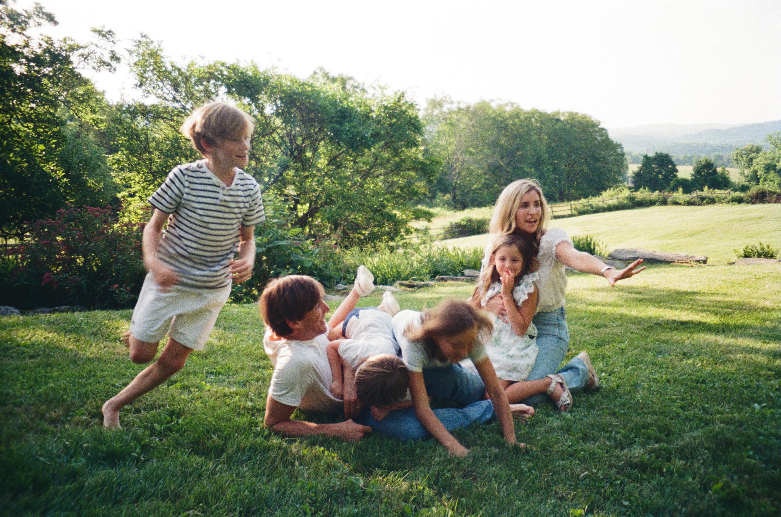 A group of children and a woman are playing and wrestling on a lush green lawn outdoors during daytime. The background features trees and rolling hills.