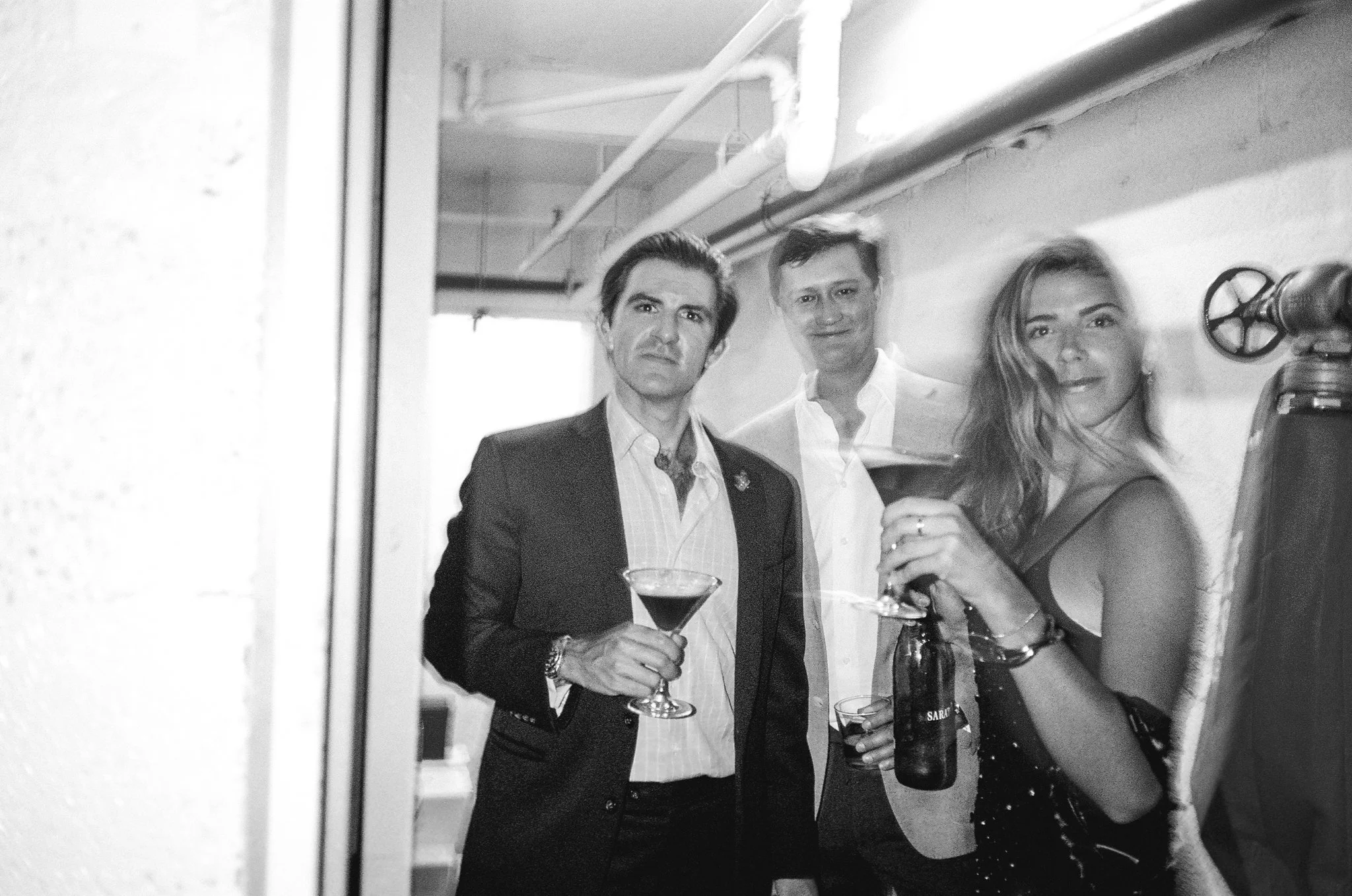 Black and white photo of three people at a social gathering, two men and one woman, holding drinks and looking at the camera in an indoor setting with pipes on the ceiling.
