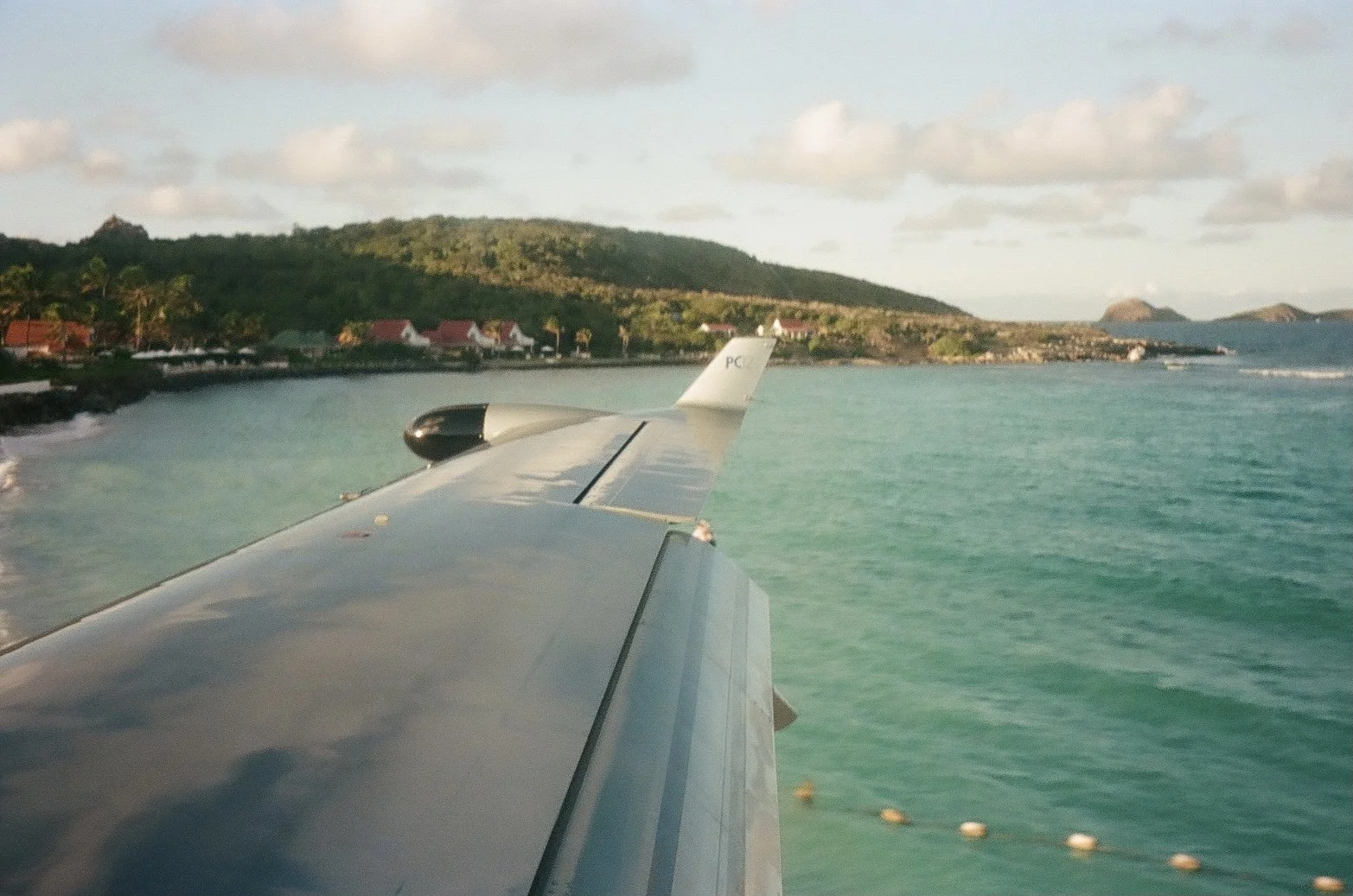 View from an airplane wing flying over a tropical island with turquoise waters and green land.