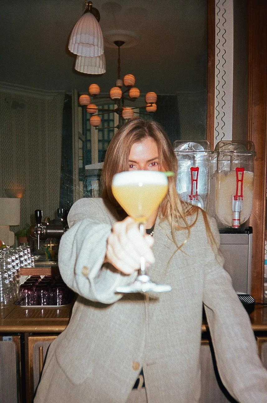 A woman holding a cocktail glass with a lime garnish, facing the camera, in a bar or restaurant setting with glasses and drink dispensers behind her.