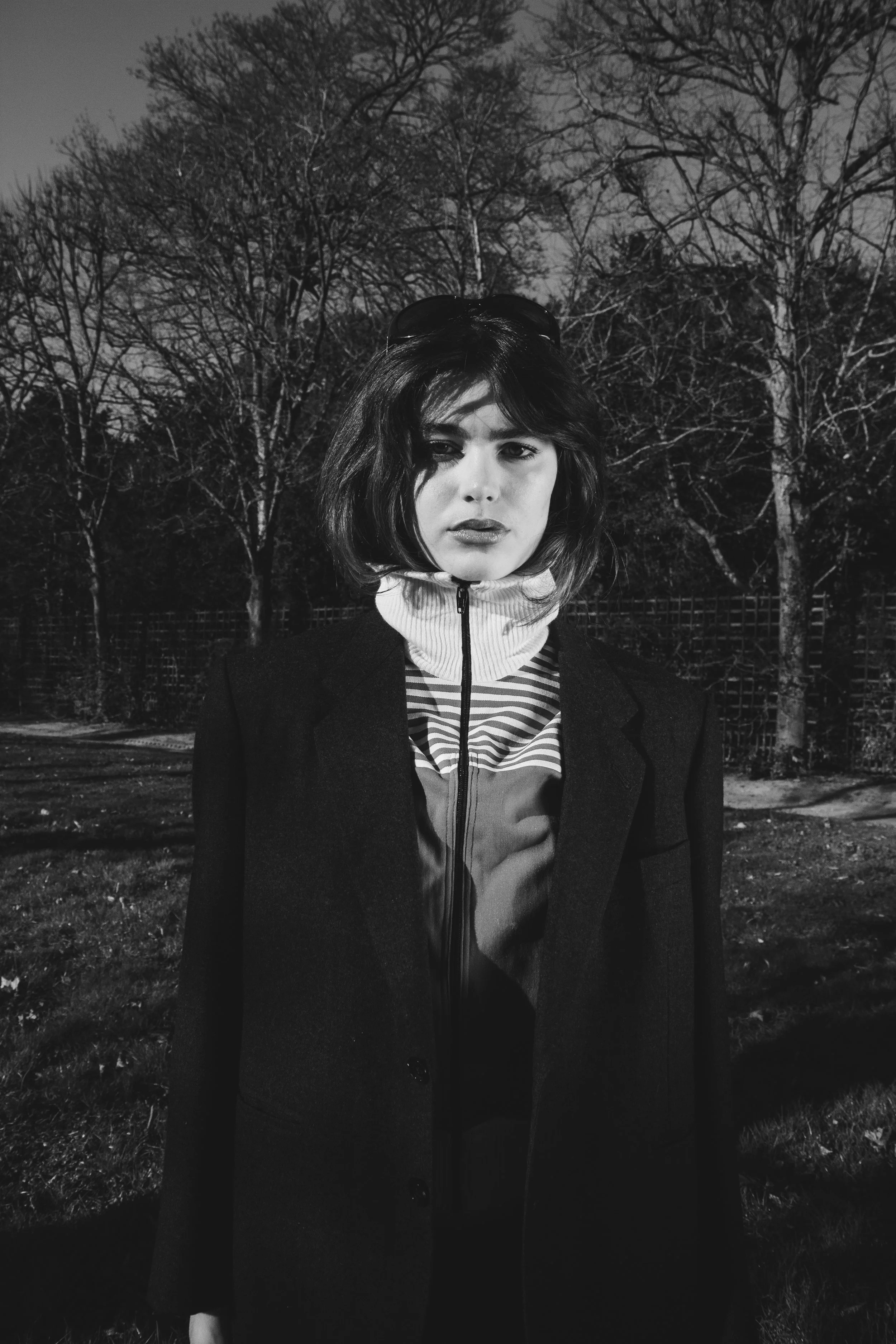 A young woman with shoulder-length wavy hair stands outdoors in front of leafless trees at night. She is wearing a dark blazer over a zip-up shirt with a striped collar and a turtleneck. The photo is in black and white.