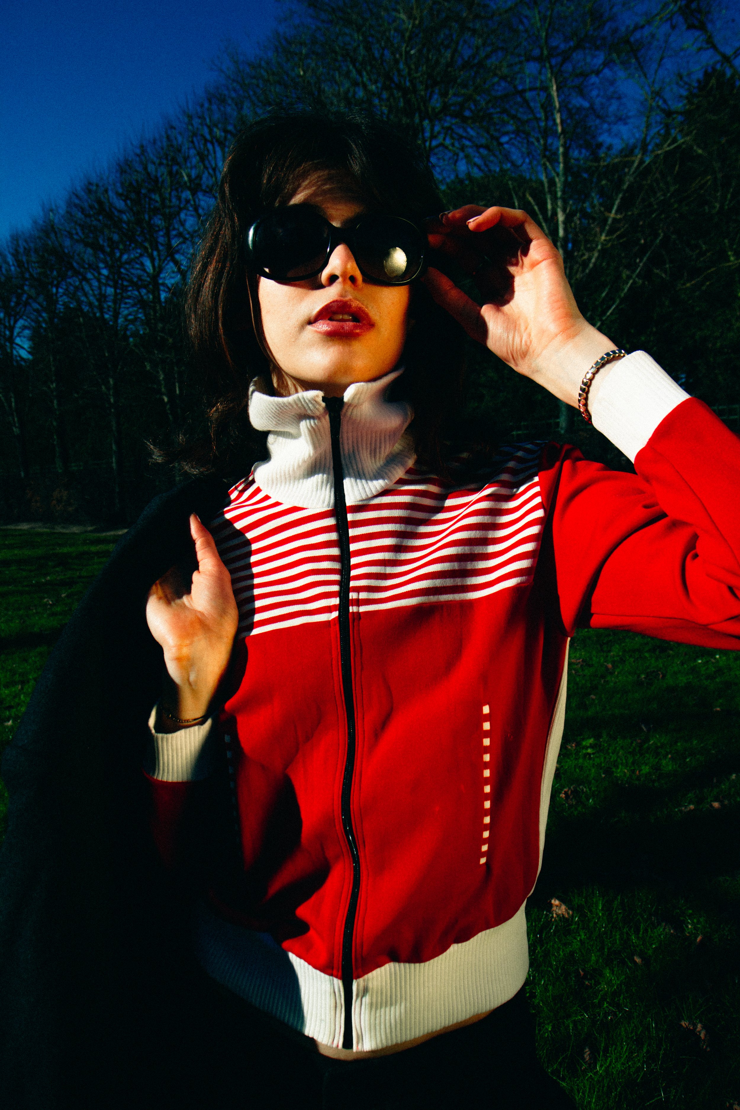 A woman wearing dark sunglasses, a red and white patterned zip-up jacket, and a white turtleneck, standing outdoors against a background of trees and a darkening sky, possibly at dusk.