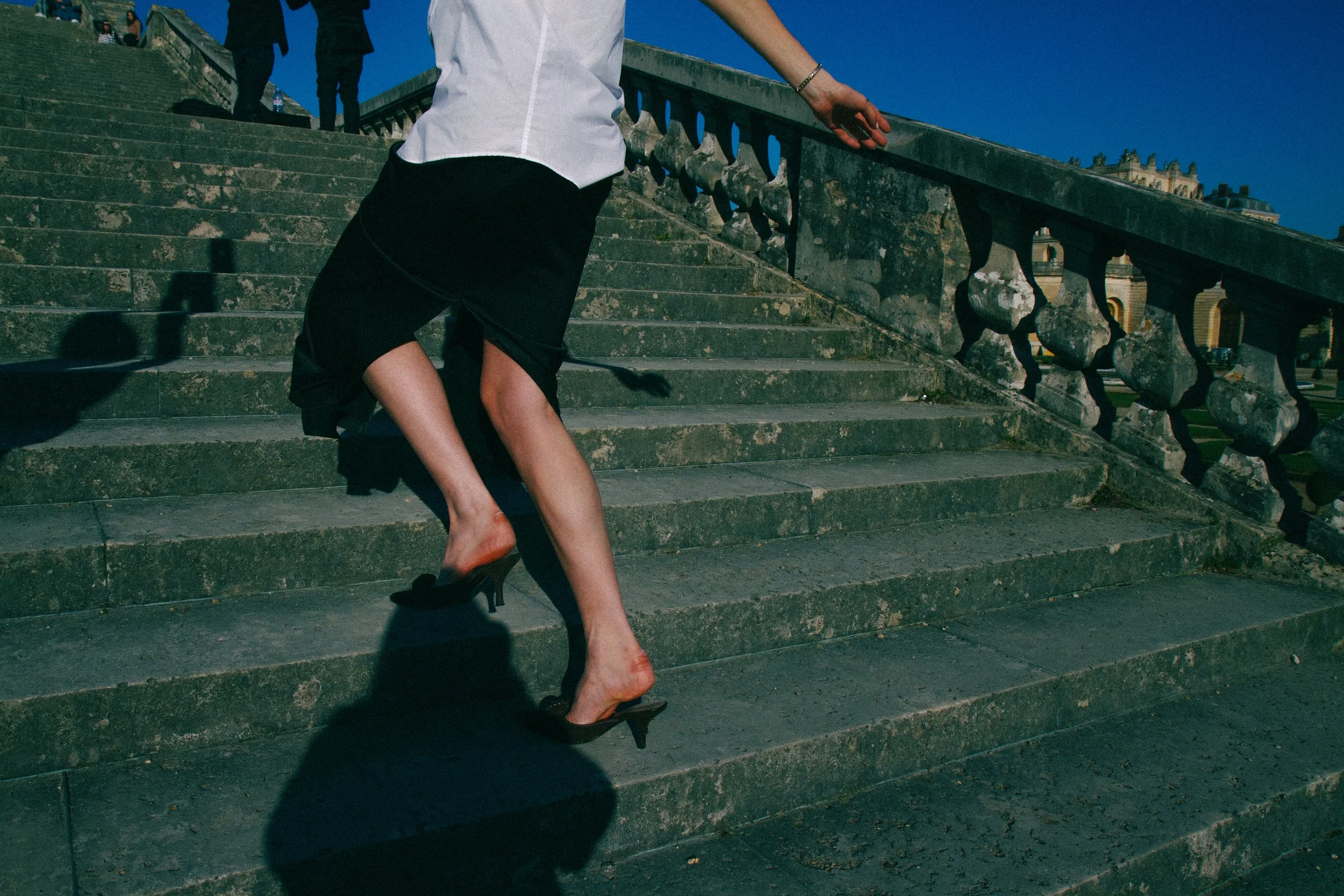 A woman wearing a black skirt, white blouse, and high heels is climbing stairs outdoors. Her face is not visible, and the photo is taken from a low angle showing her legs and part of her torso. The background shows a balustrade and a building.