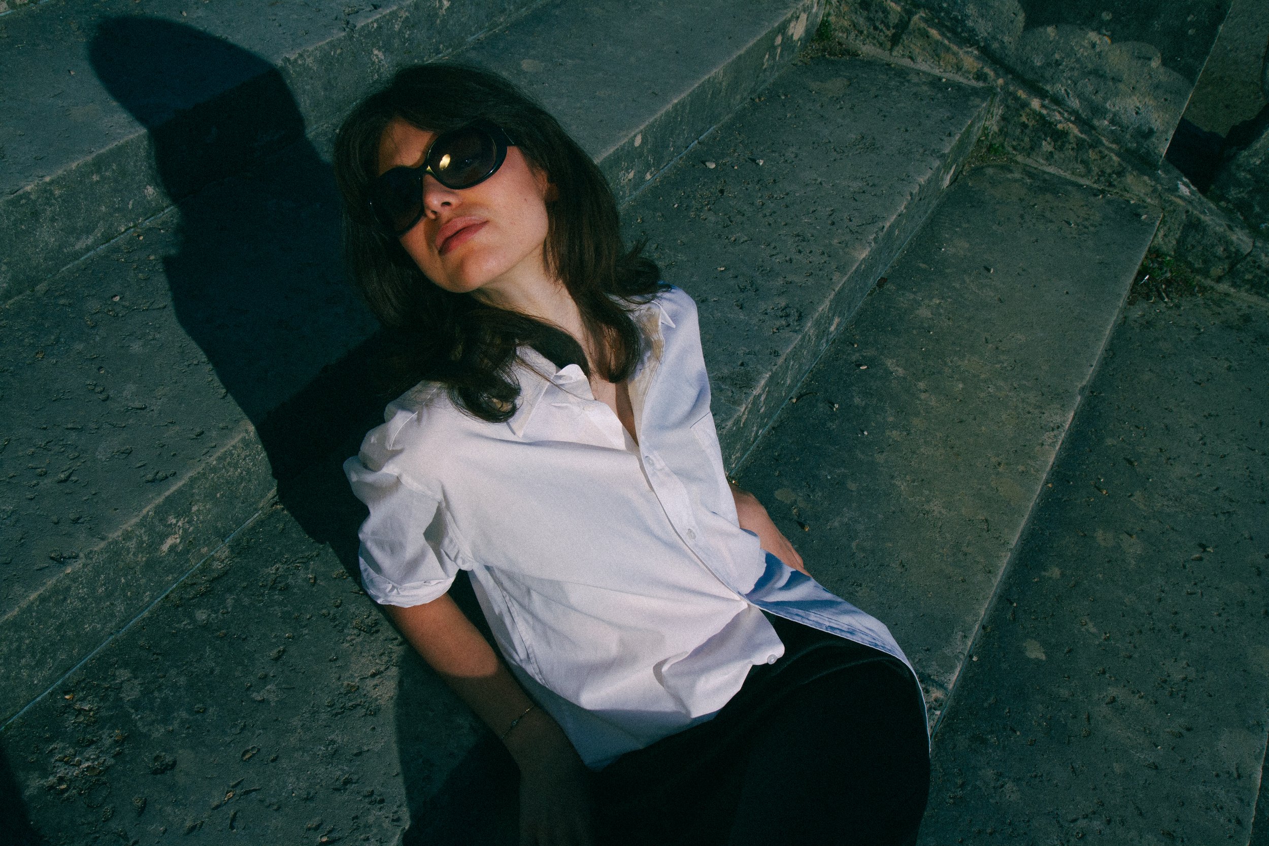 Woman with black hair and sunglasses sitting on concrete stairs, wearing a white shirt with rolled-up sleeves.
