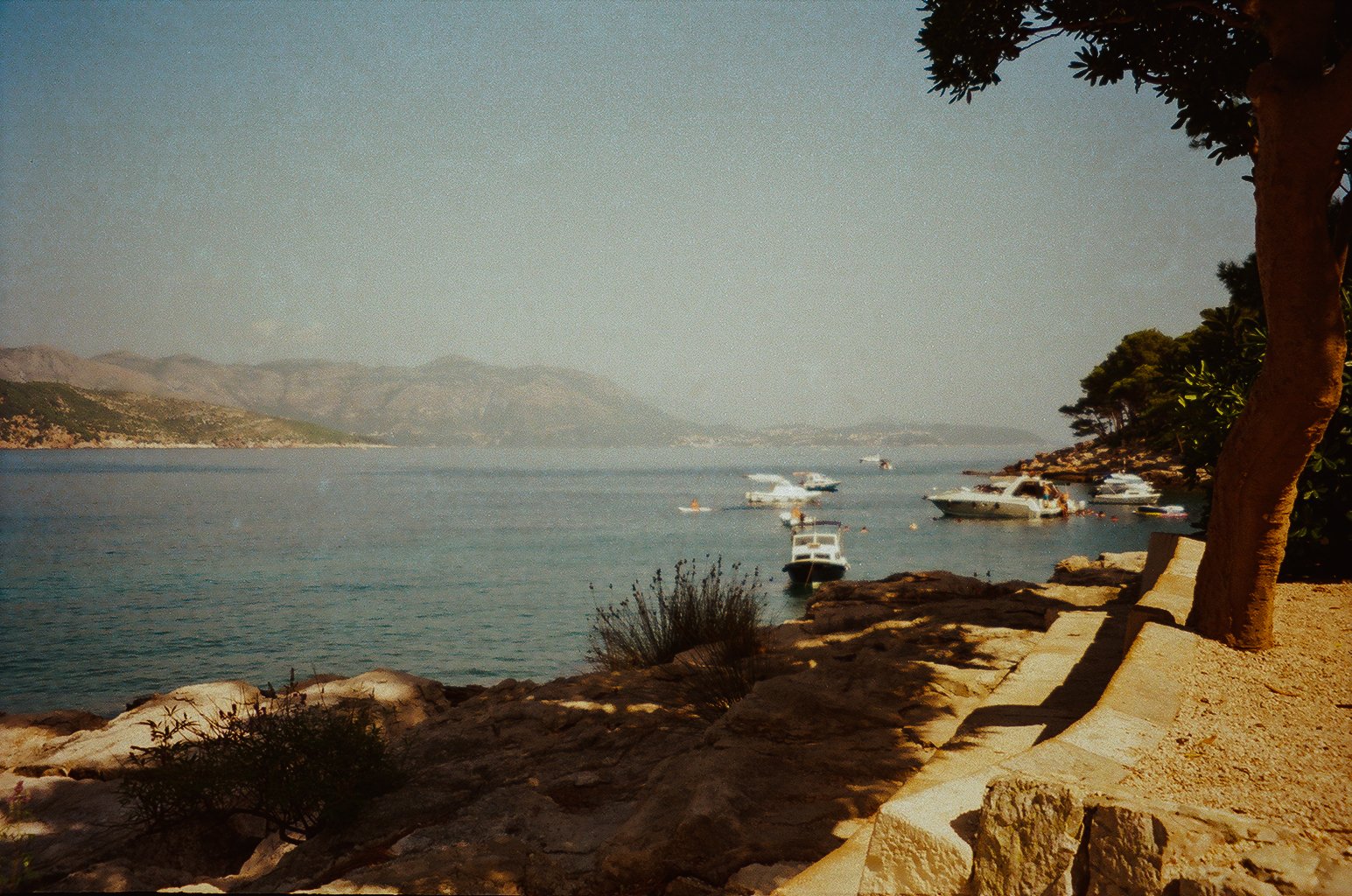 A coastal scene with boats anchored near a rocky shoreline, trees casting shadows, and a mountain range in the background.