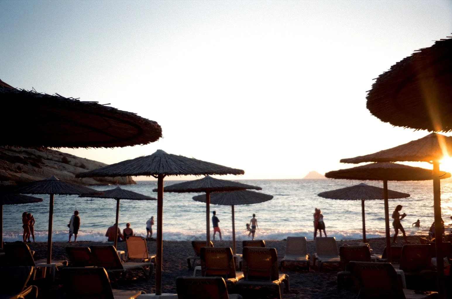 Beach scene with sun umbrellas and lounge chairs, people walking and swimming in the ocean during sunset.