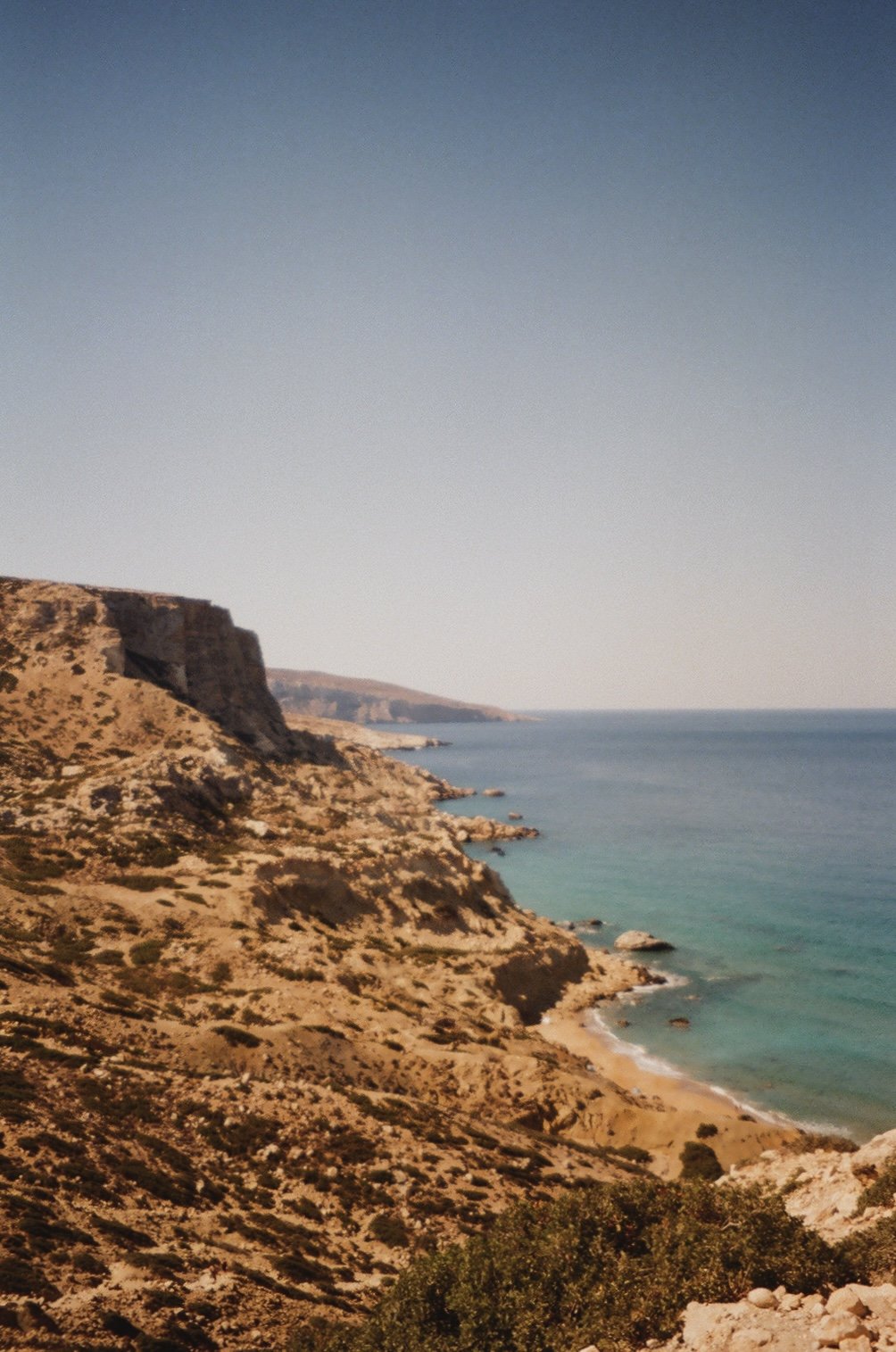 View of rocky, sandy cliffs declining to a calm ocean with a clear sky in the background.