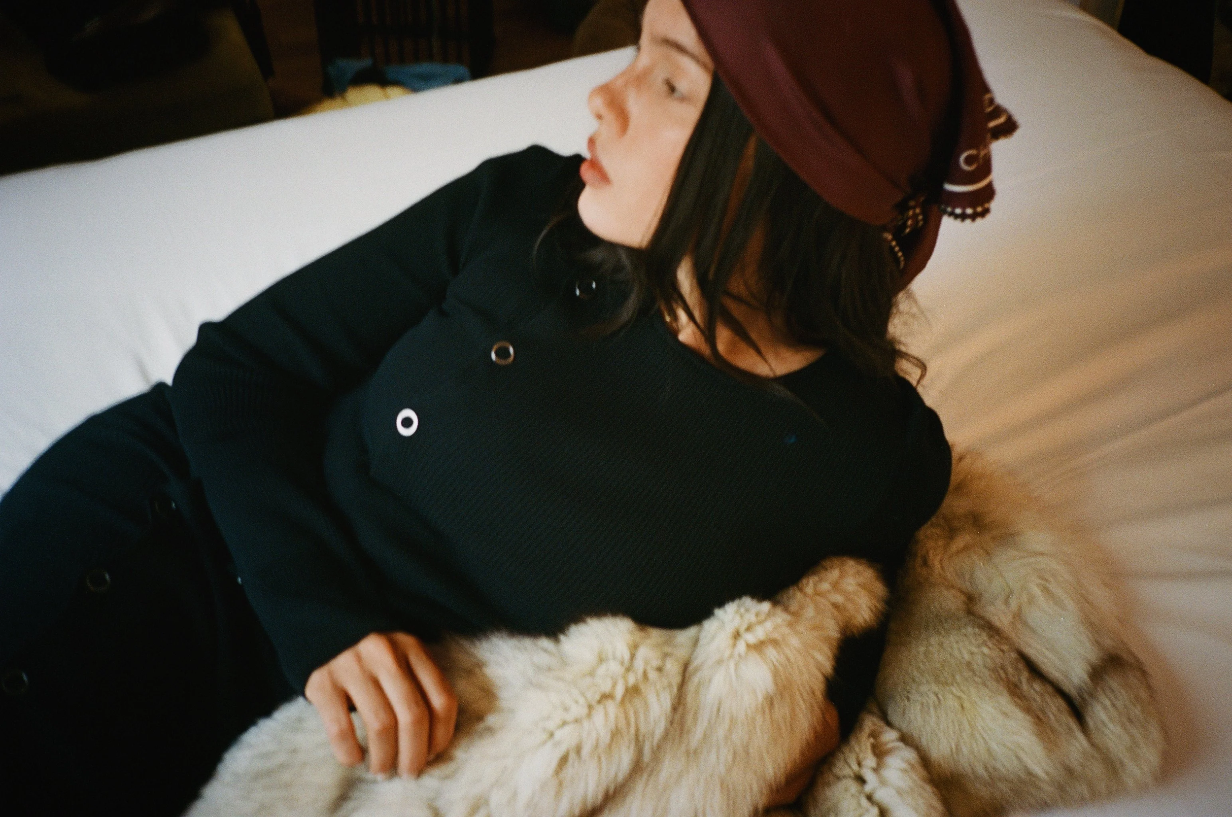 Woman lying on a bed, wearing a black jacket and a maroon headscarf, resting her head on a large, fluffy beige pillow.