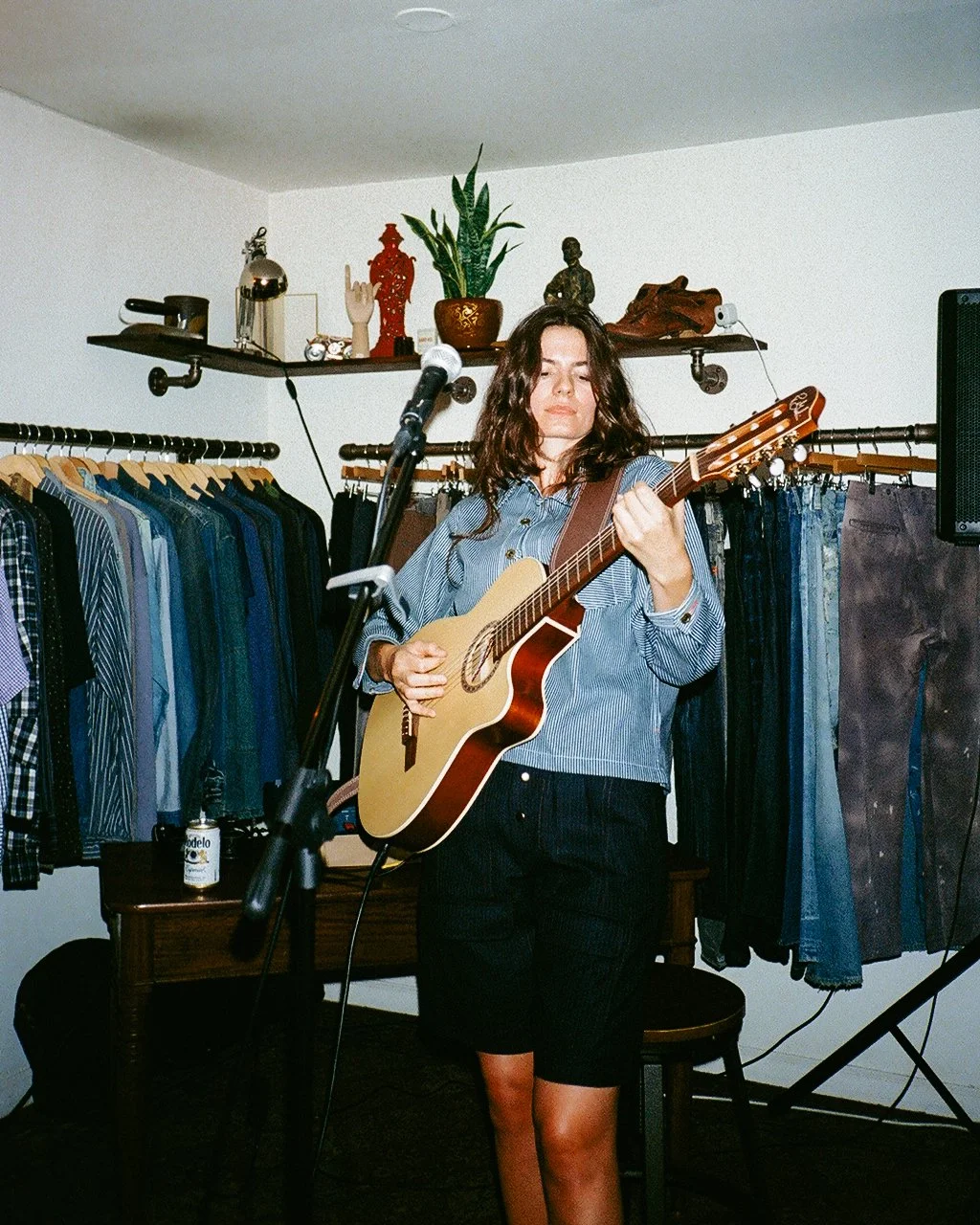 A woman with wavy brown hair playing an acoustic guitar in a clothing store or boutique. She is standing in front of a rack with various shirts and jeans, with decorations on a shelf behind her.