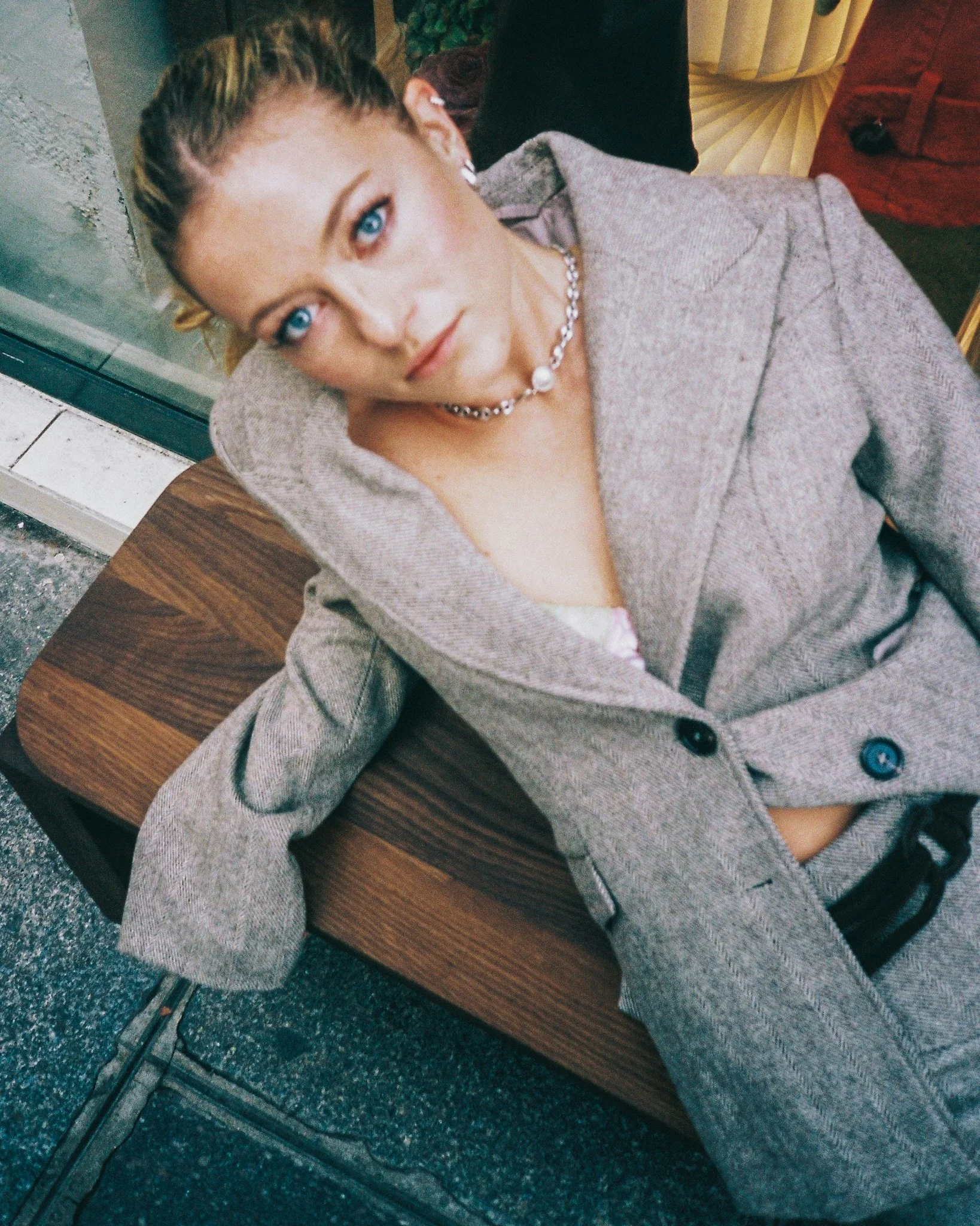 A woman with blonde hair styled in braids, blue eyes, wearing a gray blazer and a pearl necklace, leaning on a wooden table.