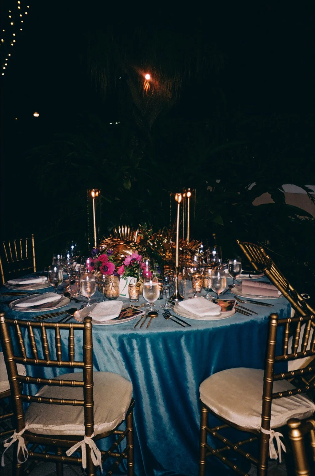A round dining table set for a special occasion with a blue tablecloth, gold chairs, floral centerpiece, tall gold candles, multiple glasses, and plates with napkins, in a dimly lit environment with decorative lighting.