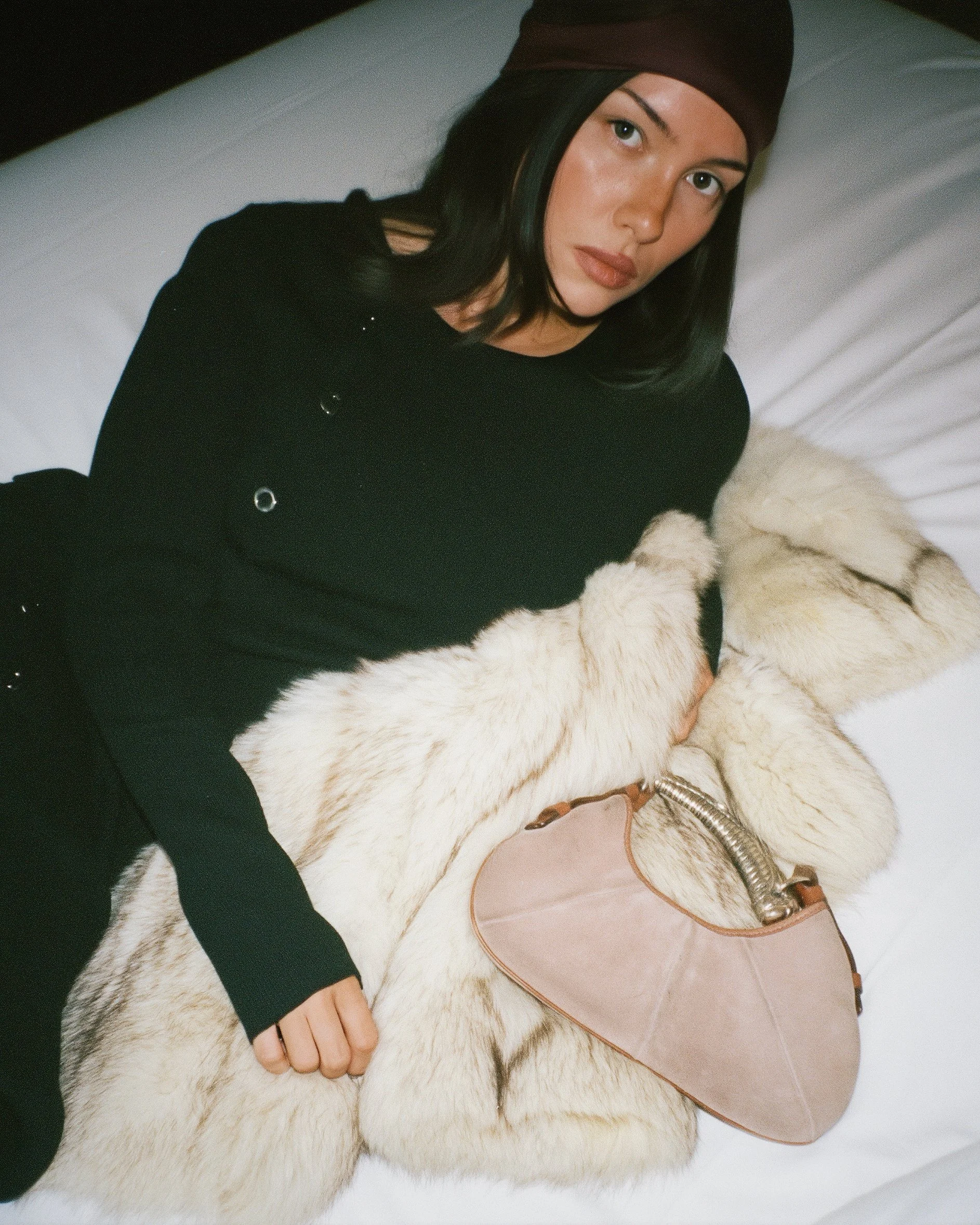 A woman with dark hair, wearing a brown headscarf, a black outfit with silver details, and beige fur gloves, is lying on a bed with white linens, holding a beige purse with a metallic handle.
