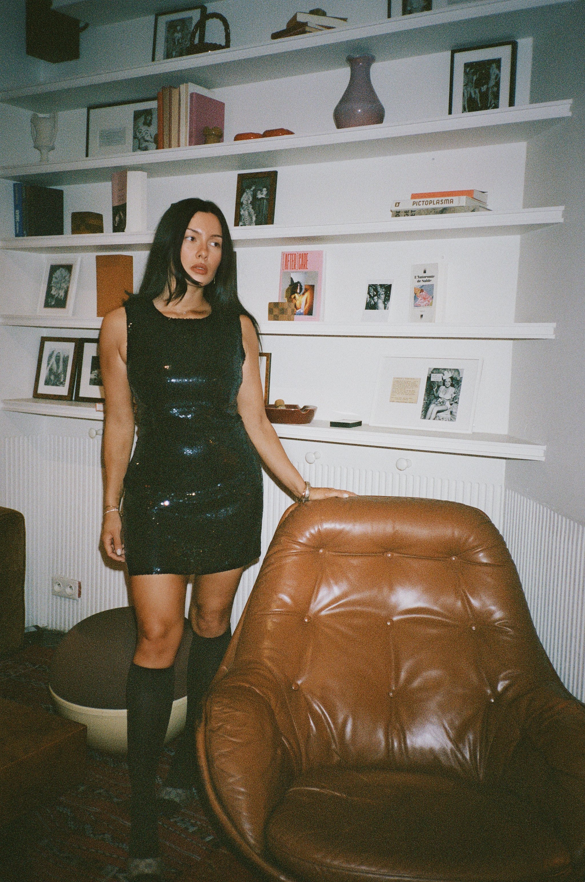 A woman in a black, sequined dress and knee-high socks standing in a room with white wall shelves decorated with framed photos and books.