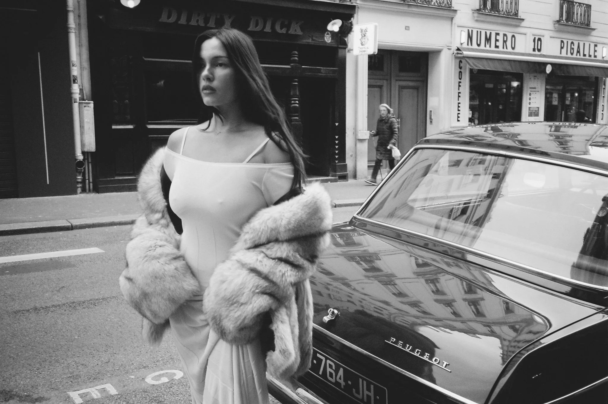 A woman in a white dress and fur coat standing on a city street near a black Peugeot car. Buildings and a pedestrian are visible in the background.