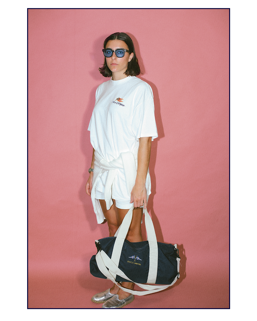 A woman wearing blue sunglasses, a white t-shirt with a small logo, white shorts with a tied shirt around her waist, silver shoes, and holding a navy blue duffel bag with white straps, standing against a pink background.