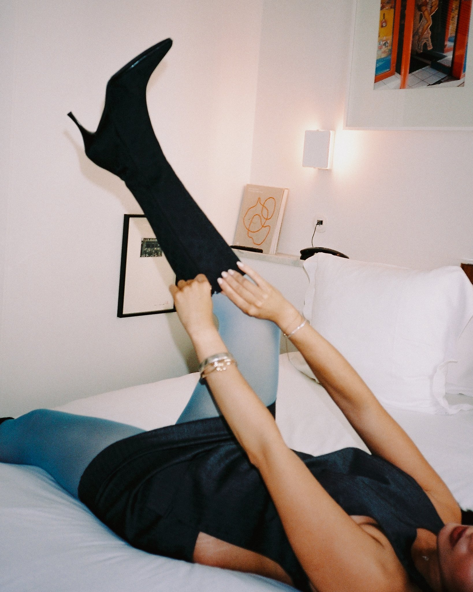 A woman lying on a bed and holding her leg up in the air, wearing a black outfit and a black high heel boot.
