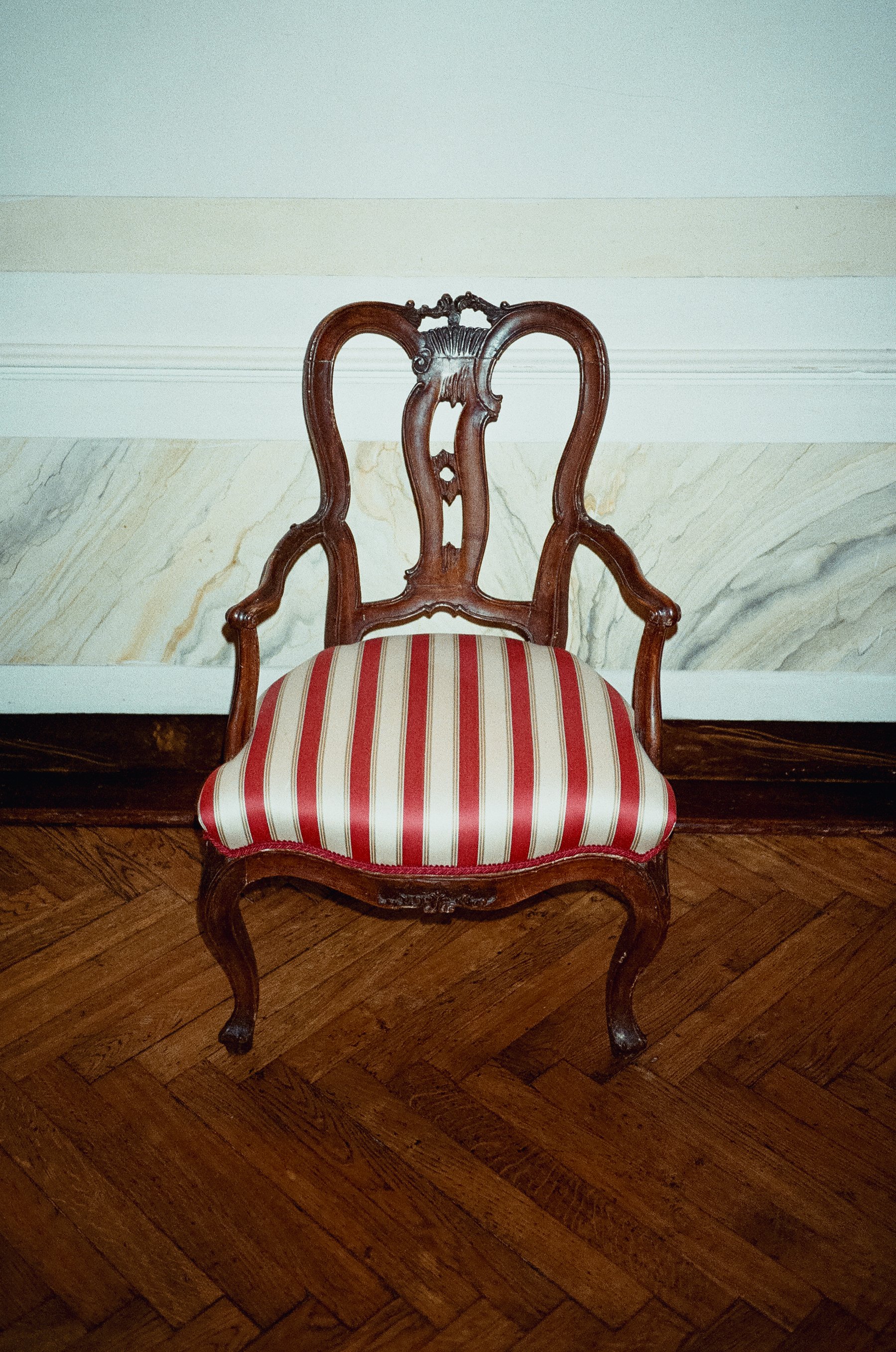 An ornate wooden chair with a striped red and cream upholstered seat, set against a wall with marble paneling and wooden flooring.