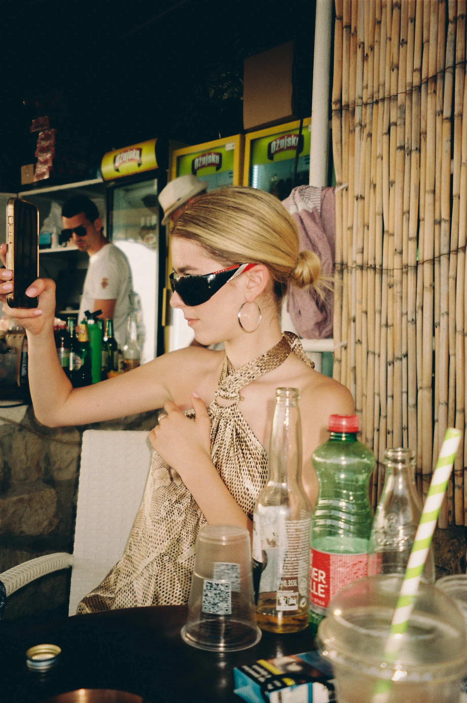 Woman with blonde hair tied in a bun, wearing sunglasses, hoop earrings, a leopard print top, and a snake necklace, sitting at a table with beverage bottles, cups, and a straw, taking a selfie at an outdoor bar or restaurant.