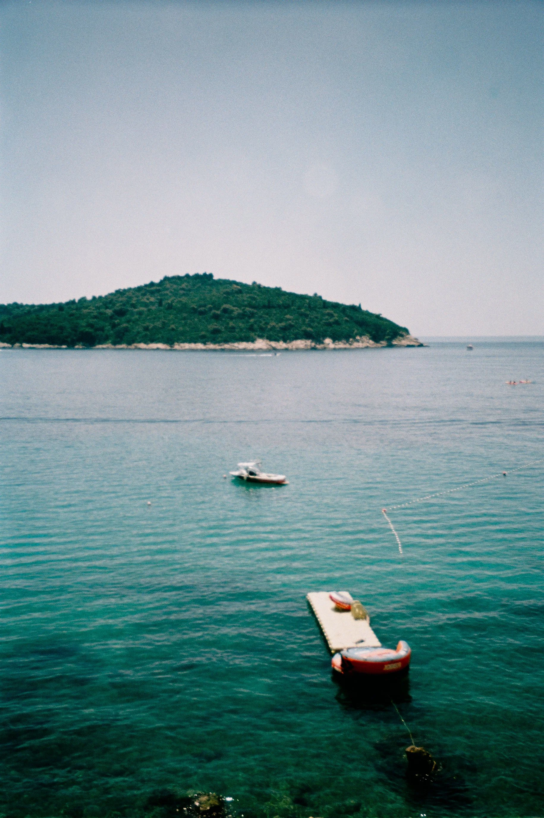 Calm sea with two boats and a floating dock near a green island in the distance, under a clear sky.