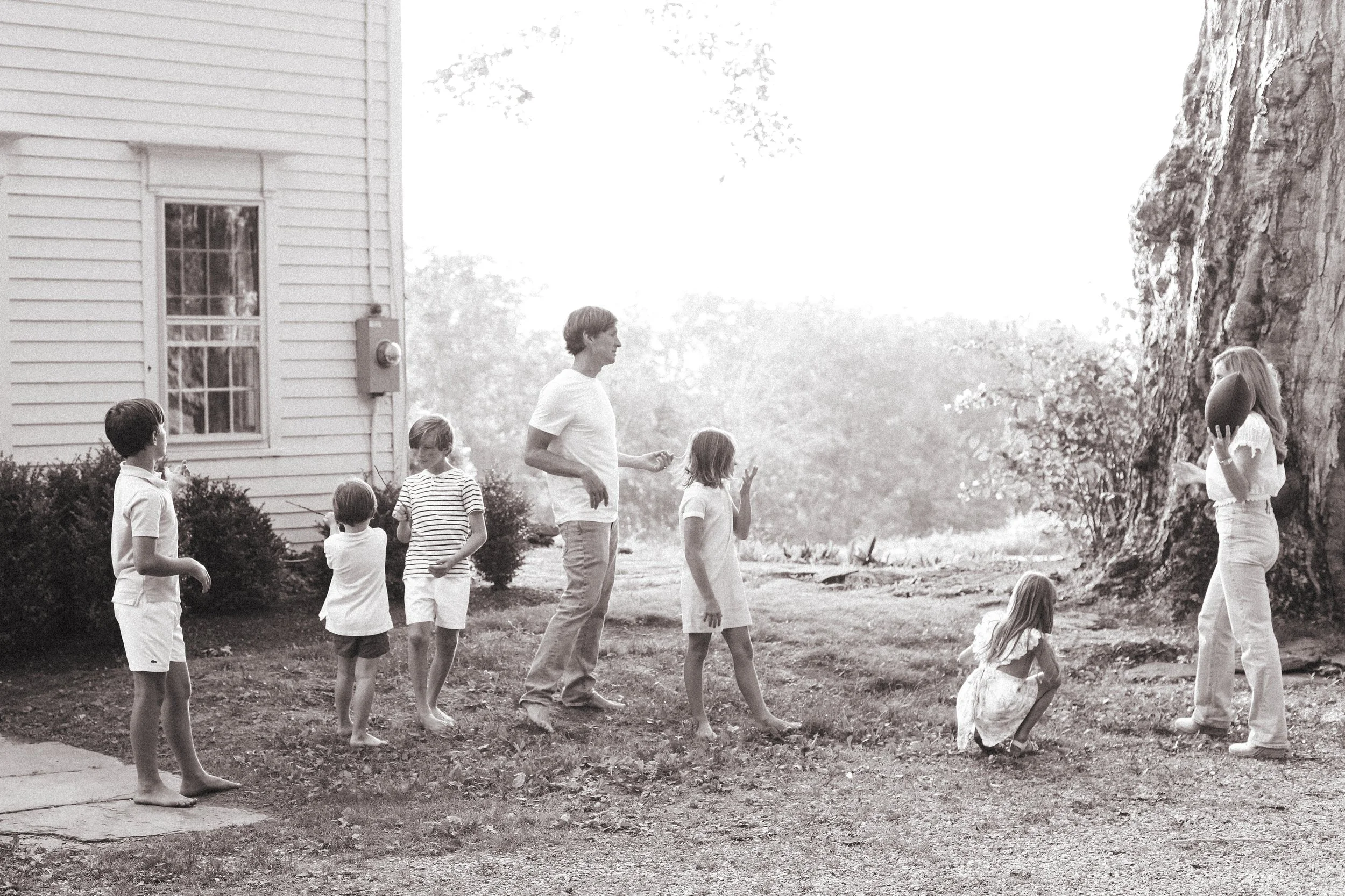 Children and an adult playing outside near a house and large tree, with a girl holding a football and another girl in a dress squatting on the ground.