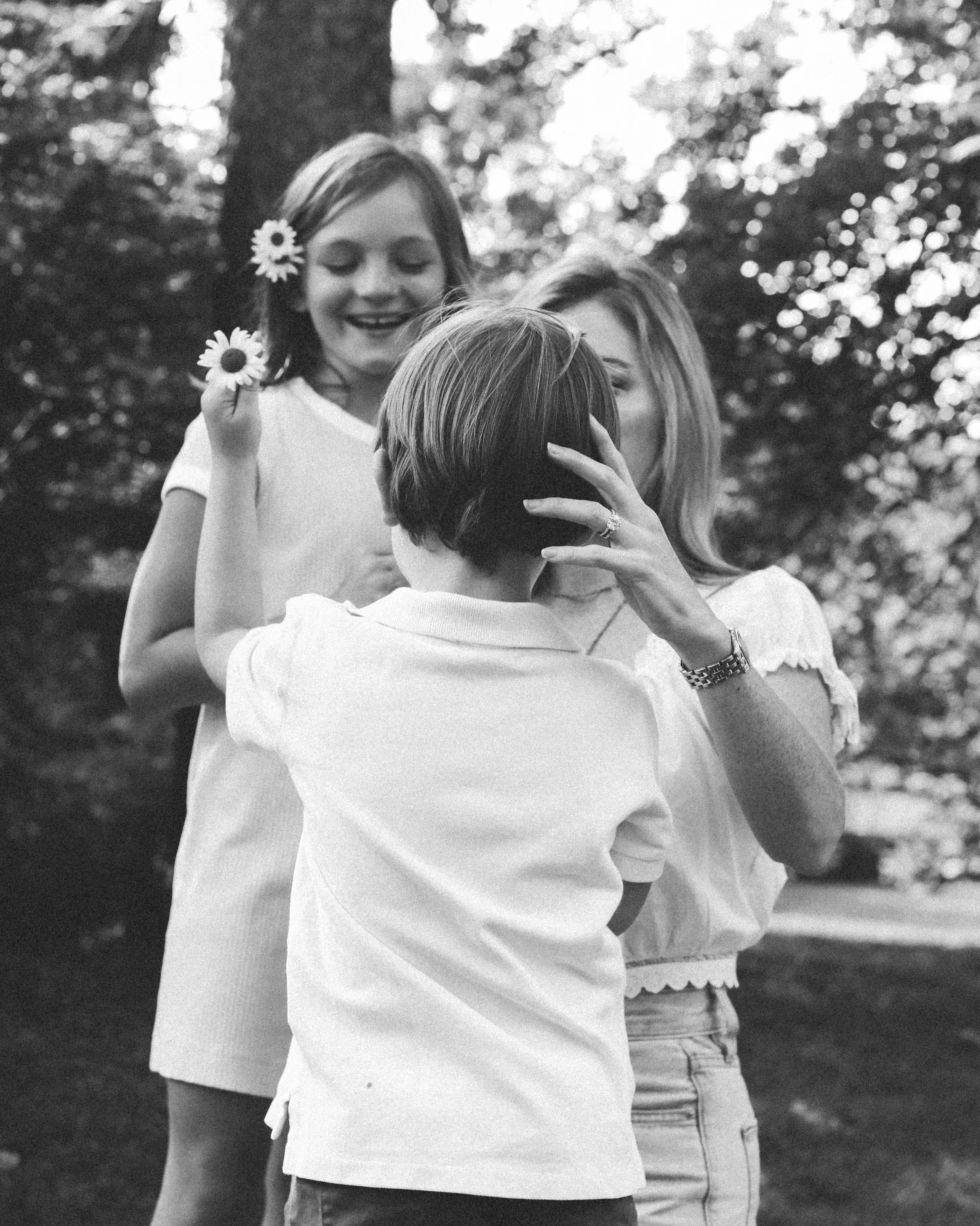 A woman is hugging a young boy, while a girl holds daisies in the background, outdoors in a wooded area.
