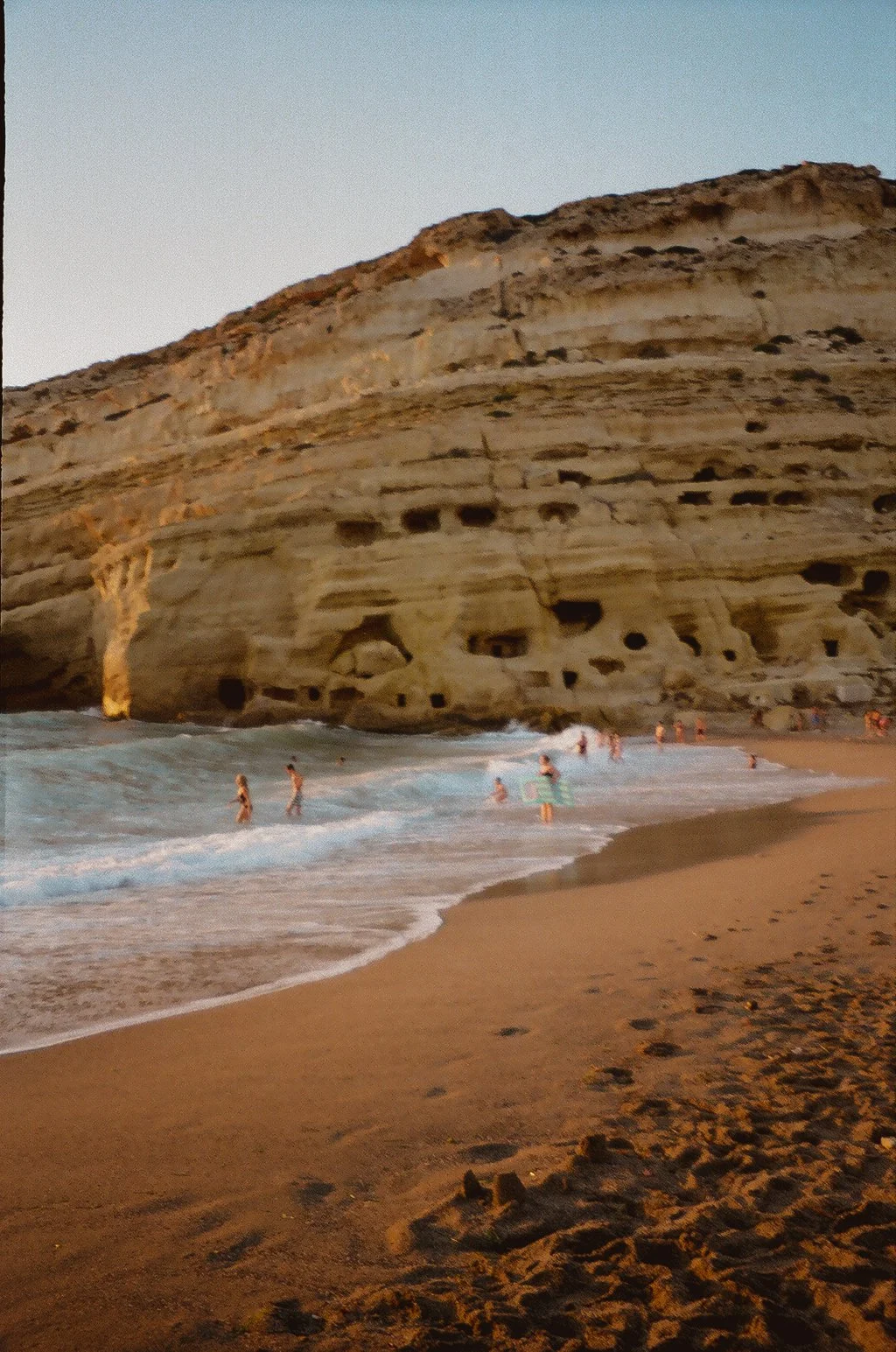 People swimming and playing in the ocean near a sandy beach with a large rocky cliff in the background during sunset.