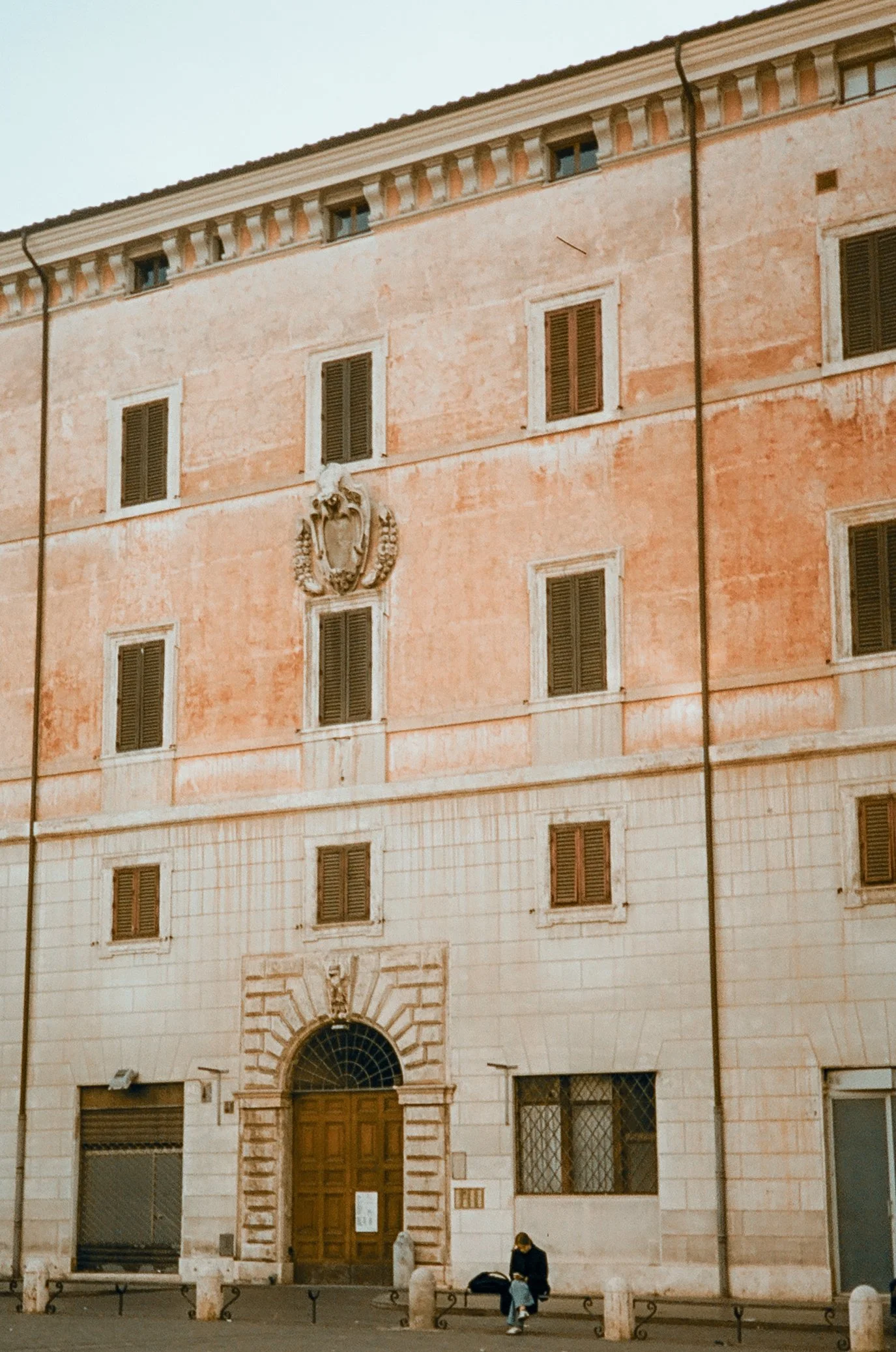Facade of a historic European building with multiple windows, an arched entrance, and decorative crest, with a person sitting on a bench outside.