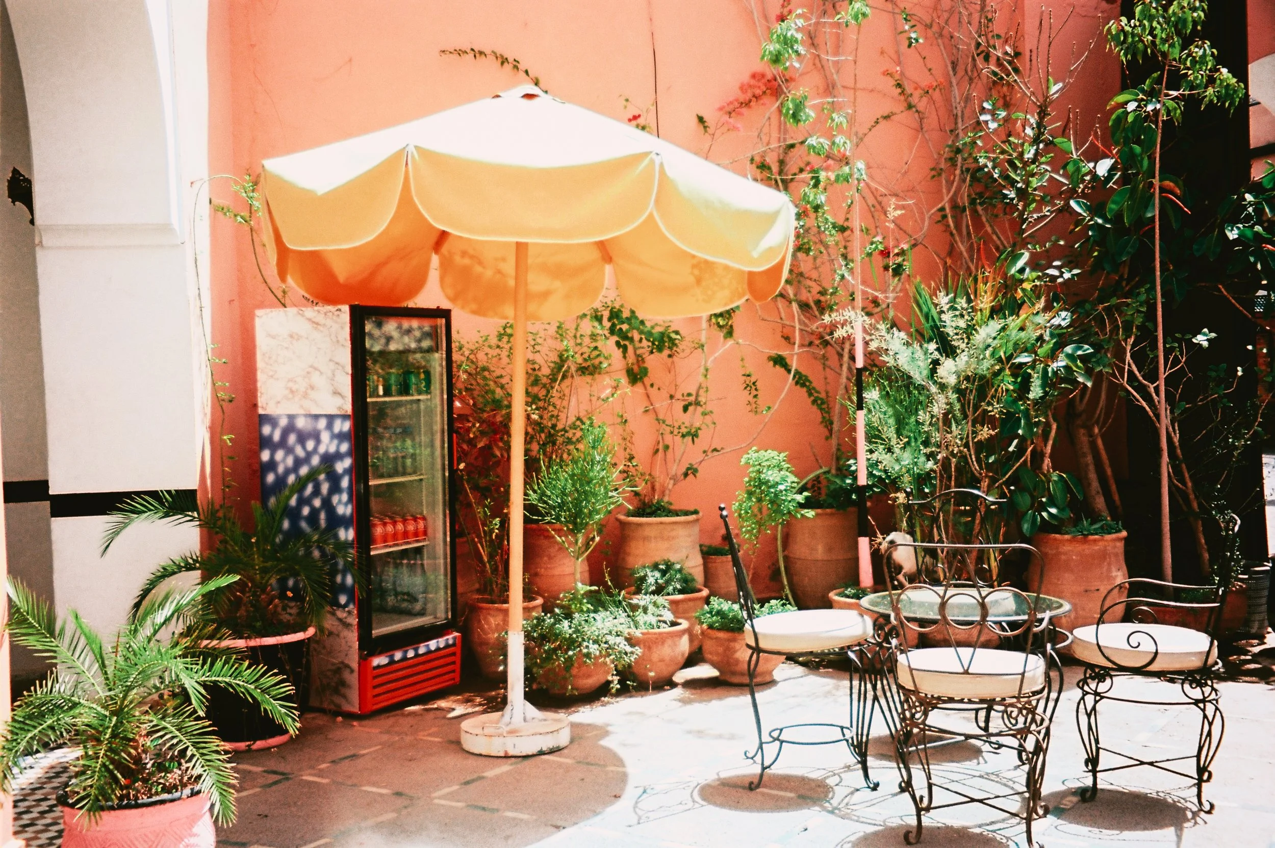 Outdoor patio with potted plants, a yellow umbrella, metal chairs and table, and a small refrigerator against a pink wall.