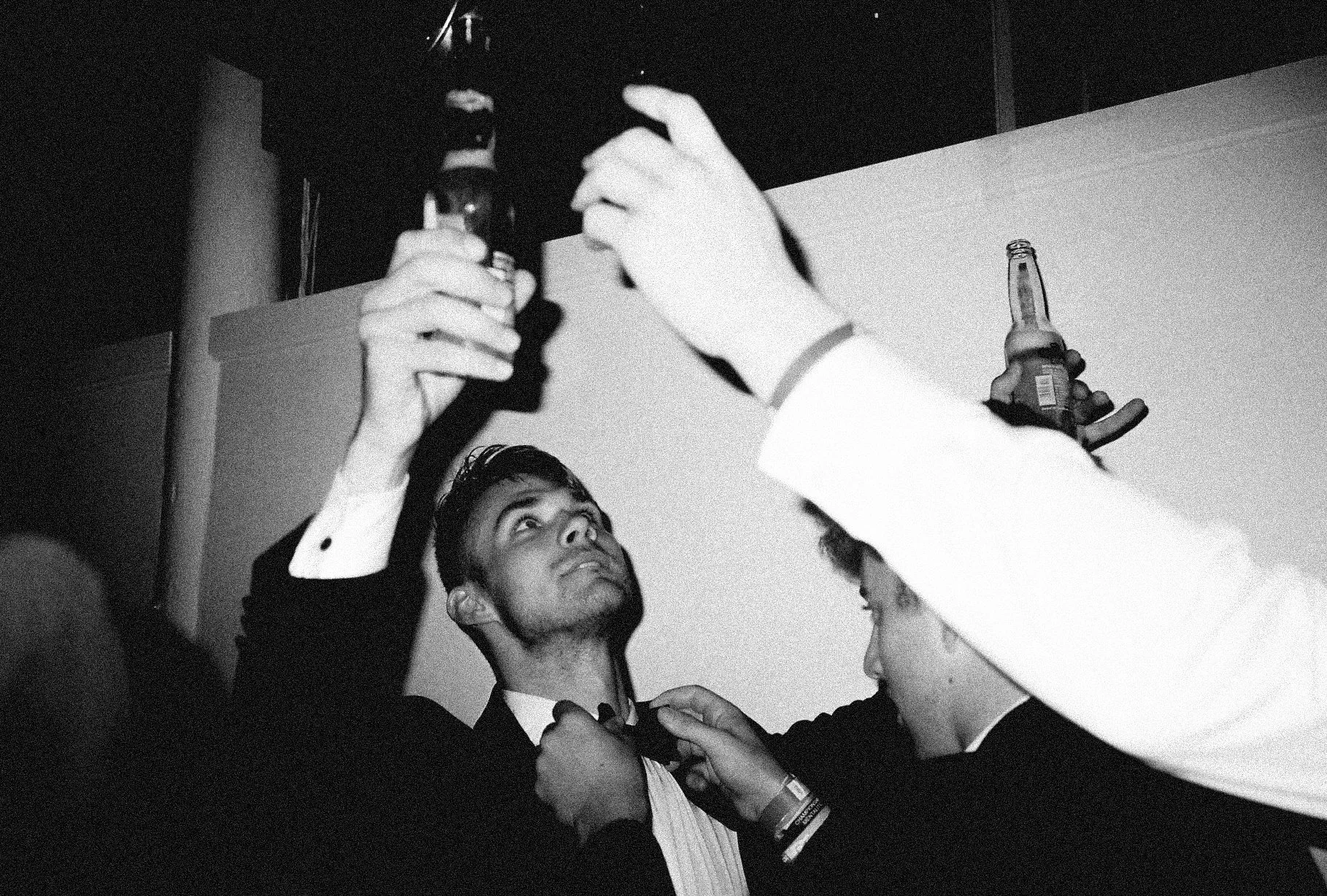 Young man in a tuxedo raises a beer bottle at a party while another person helps him with his bowtie. The scene appears lively and celebratory.