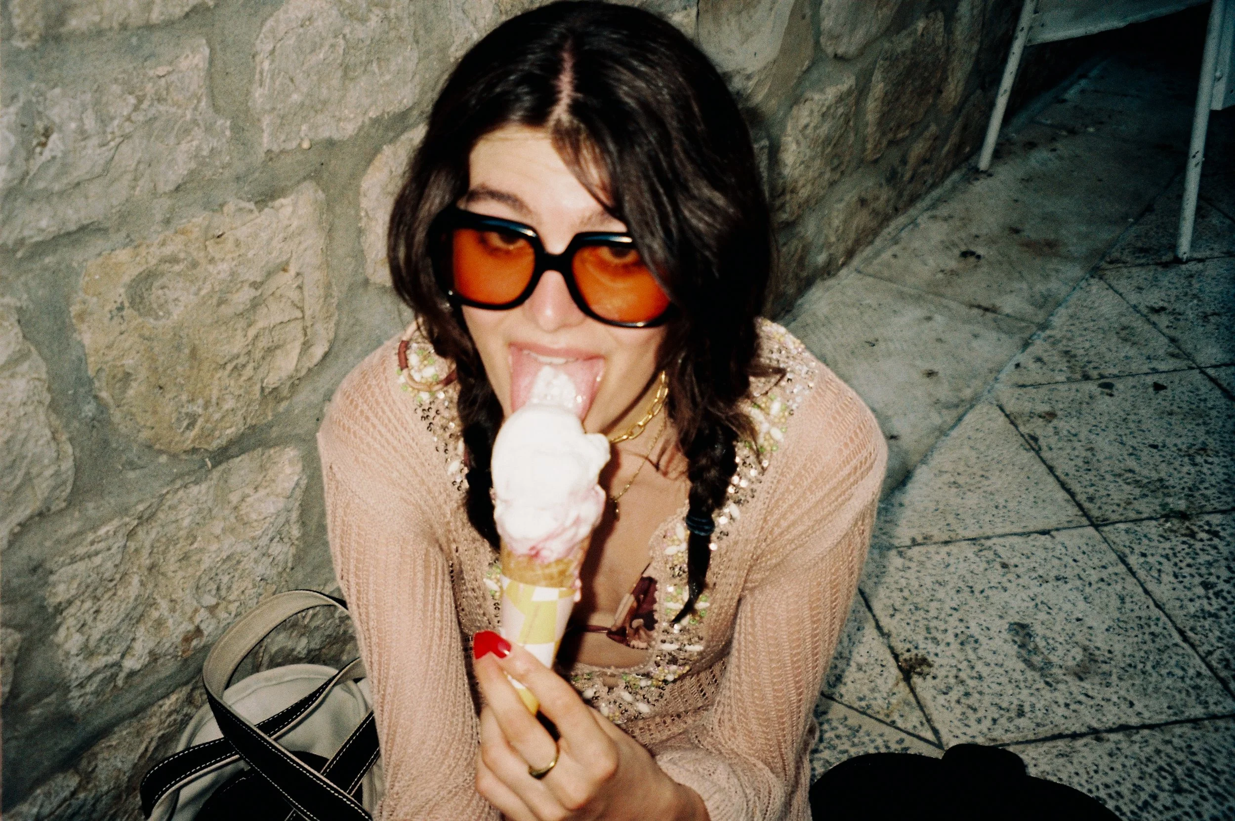 A woman with long dark hair, wearing large orange-tinted sunglasses and a beige sweater with embellishments, is sitting on the ground beside a stone wall. She is licking a vanilla ice cream cone and holding it in her right hand.