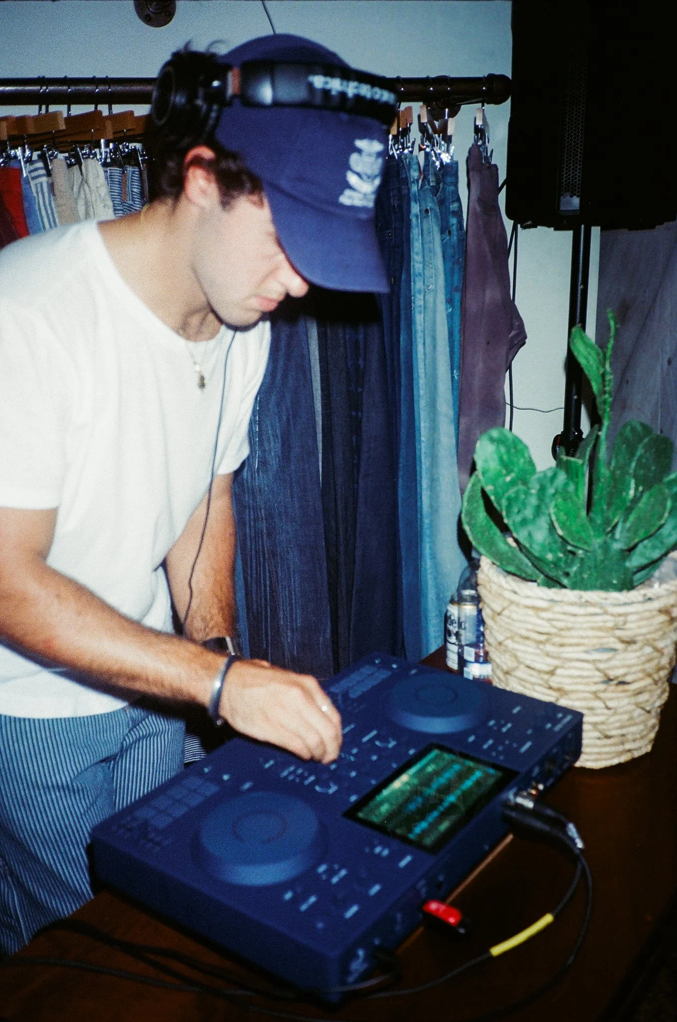 A young man in a white t-shirt and striped pants using DJ equipment with a headset on. Behind him is clothing hanging on a rack and a large potted plant on a table.