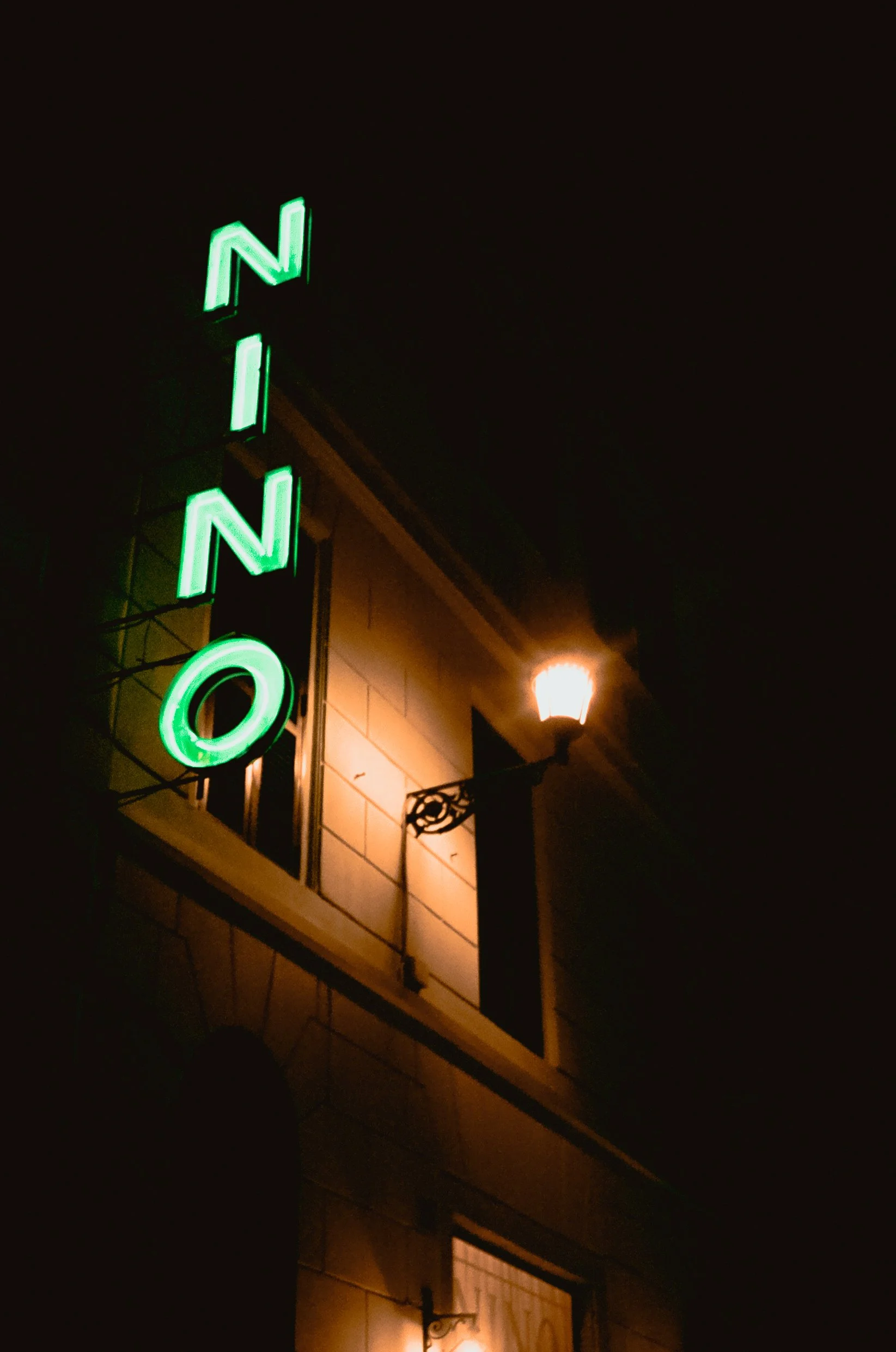 Nighttime view of a building with a green neon sign spelling 'NINO' and a lit outdoor lamp.