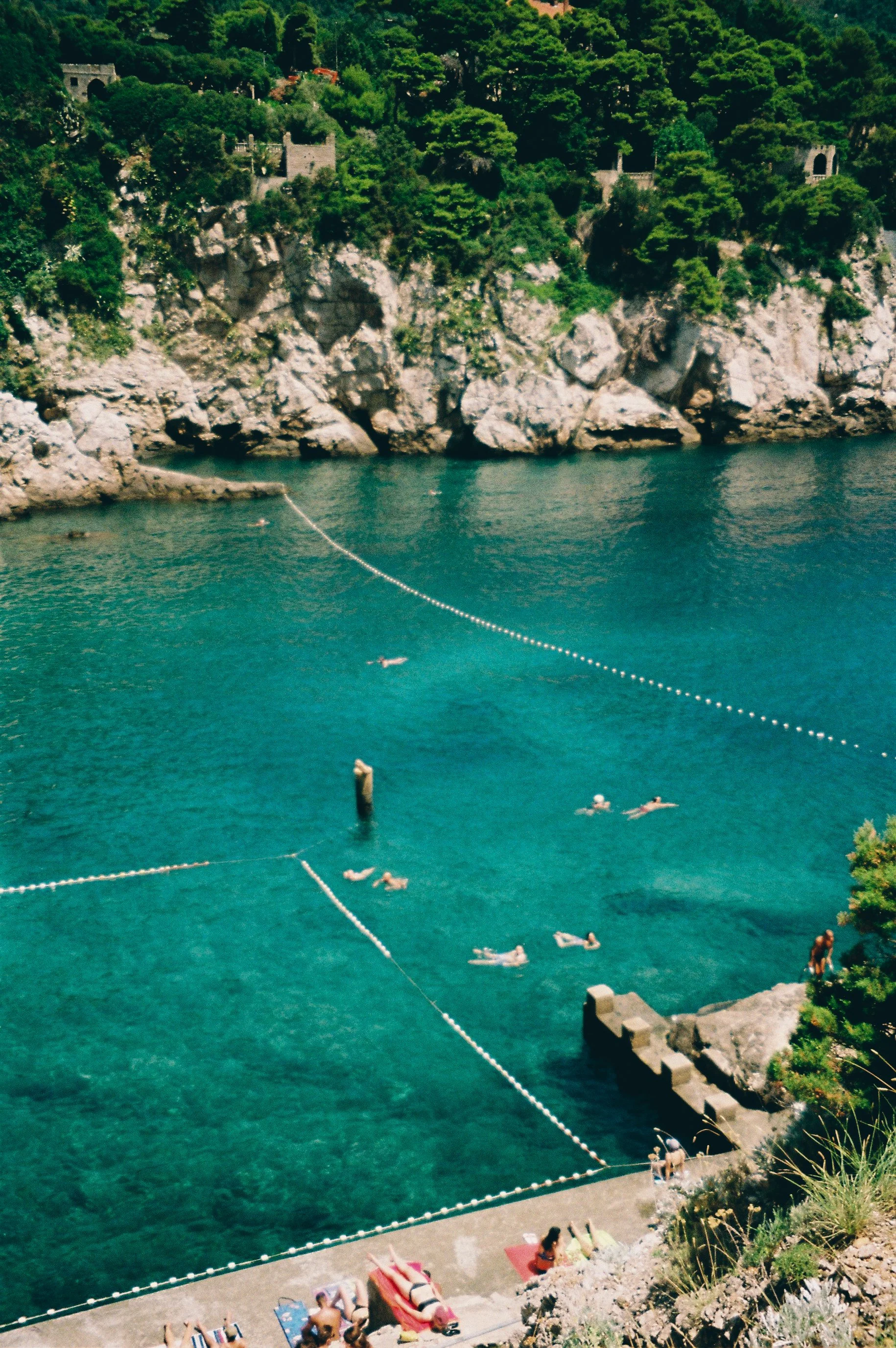 People swimming and relaxing at a seaside swimming area with a concrete dock, rocky cliffs, and lush green trees in the background.