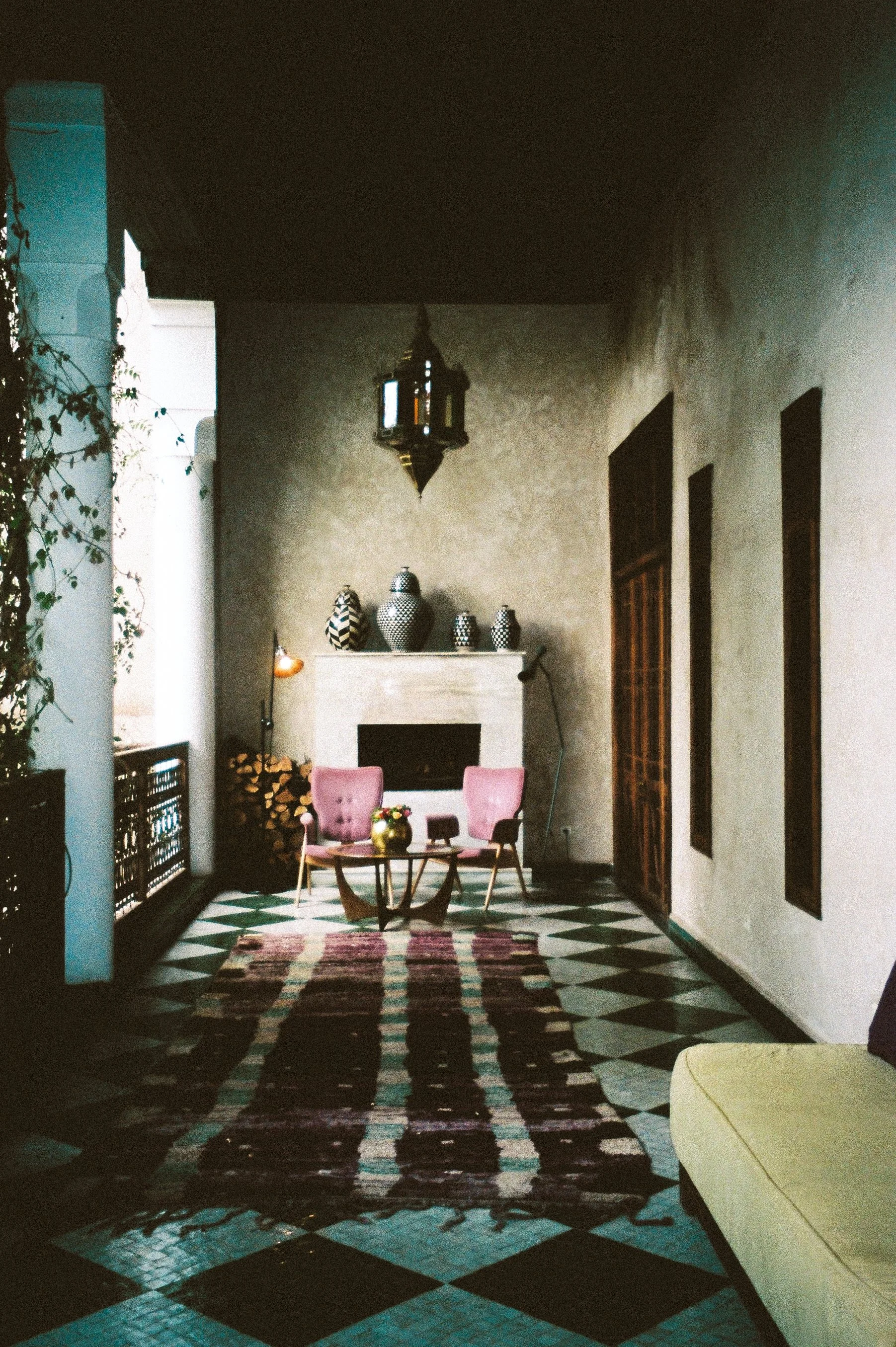 Interior view of a cozy sitting area with pink armchairs, a patterned rug, a fireplace decorated with vases, a hanging lantern, and a yellow sofa on the right.