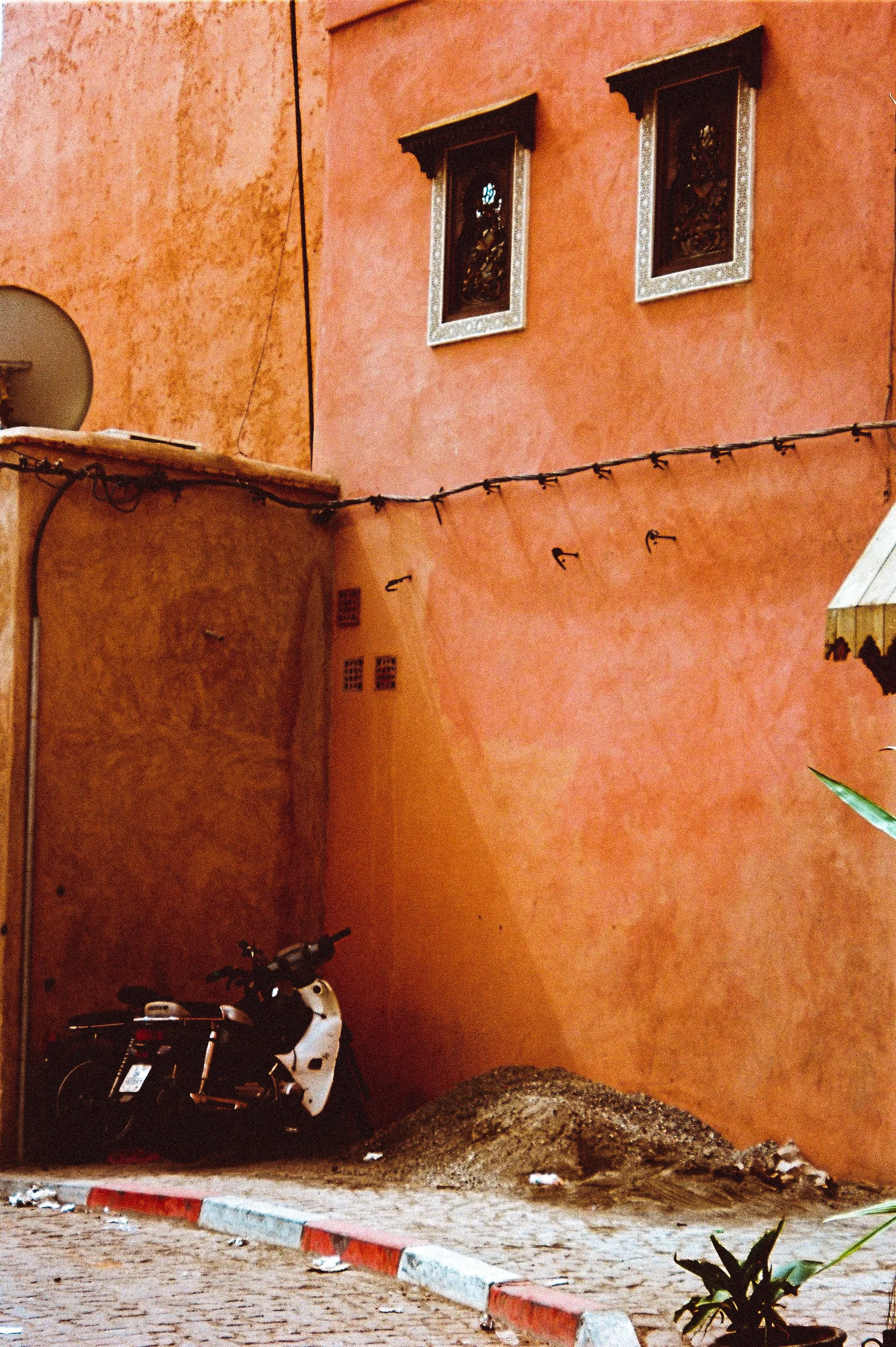 A corner of a cobblestone street with a red and white painted curb, featuring a black and white motorcycle parked next to a peach-colored wall of a building. The building has two small windows with ornate frames, and a string of lights is hanging acr