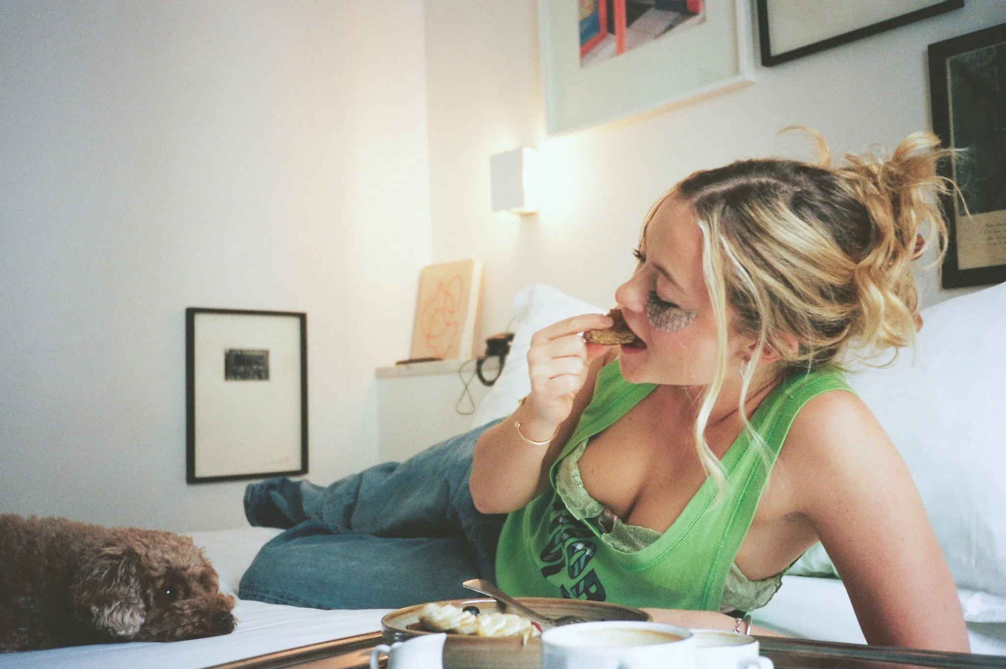 A woman with curly blonde hair, wearing a green tank top, is lying on a bed and eating a snack while petting a small brown dog nearby. There are framed pictures on the wall behind her and a tray with food and a cup on the bed.
