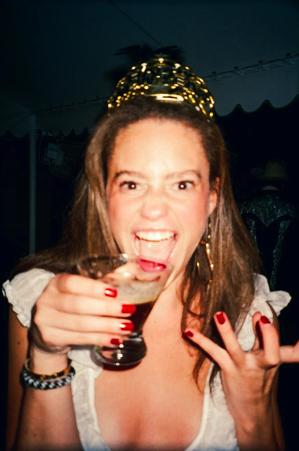 A woman with long brown hair, wearing a gold headband, holding a cocktail glass, and smiling with her mouth open. She has red nail polish and jewelry, in a festive or party setting.