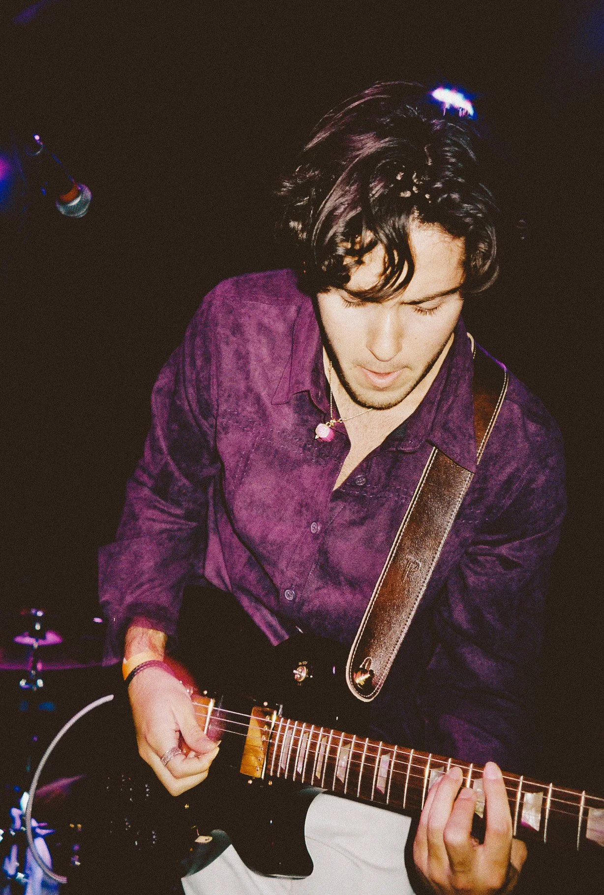 A young man with dark hair playing an electric guitar, wearing a purple shirt, on stage with dark background and stage lights.