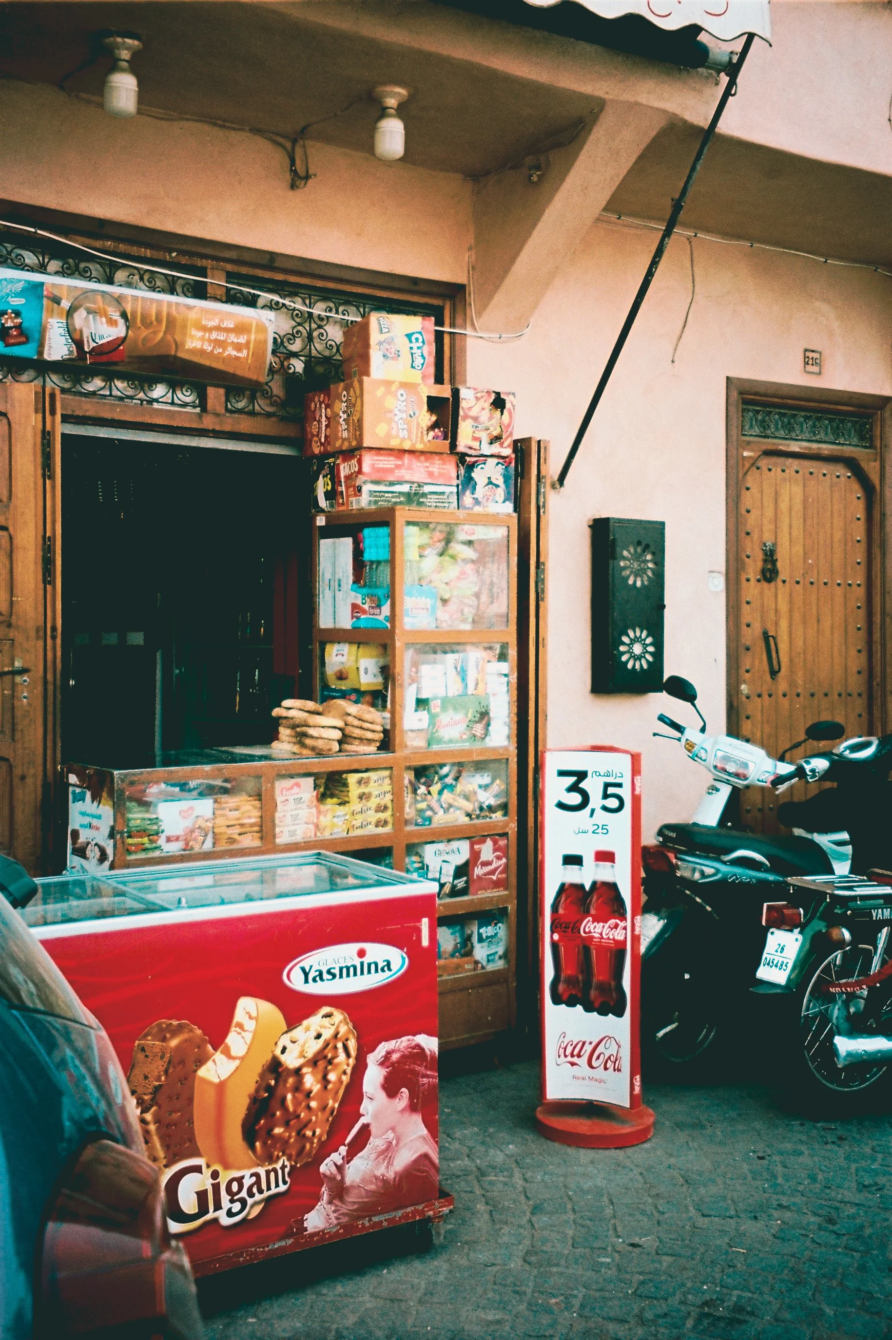 Street scene featuring a small shop with various snack boxes, a Coca-Cola ice cooler, a motorbike, and a sign advertising Coca-Cola bottles priced at 3.5 in local currency.