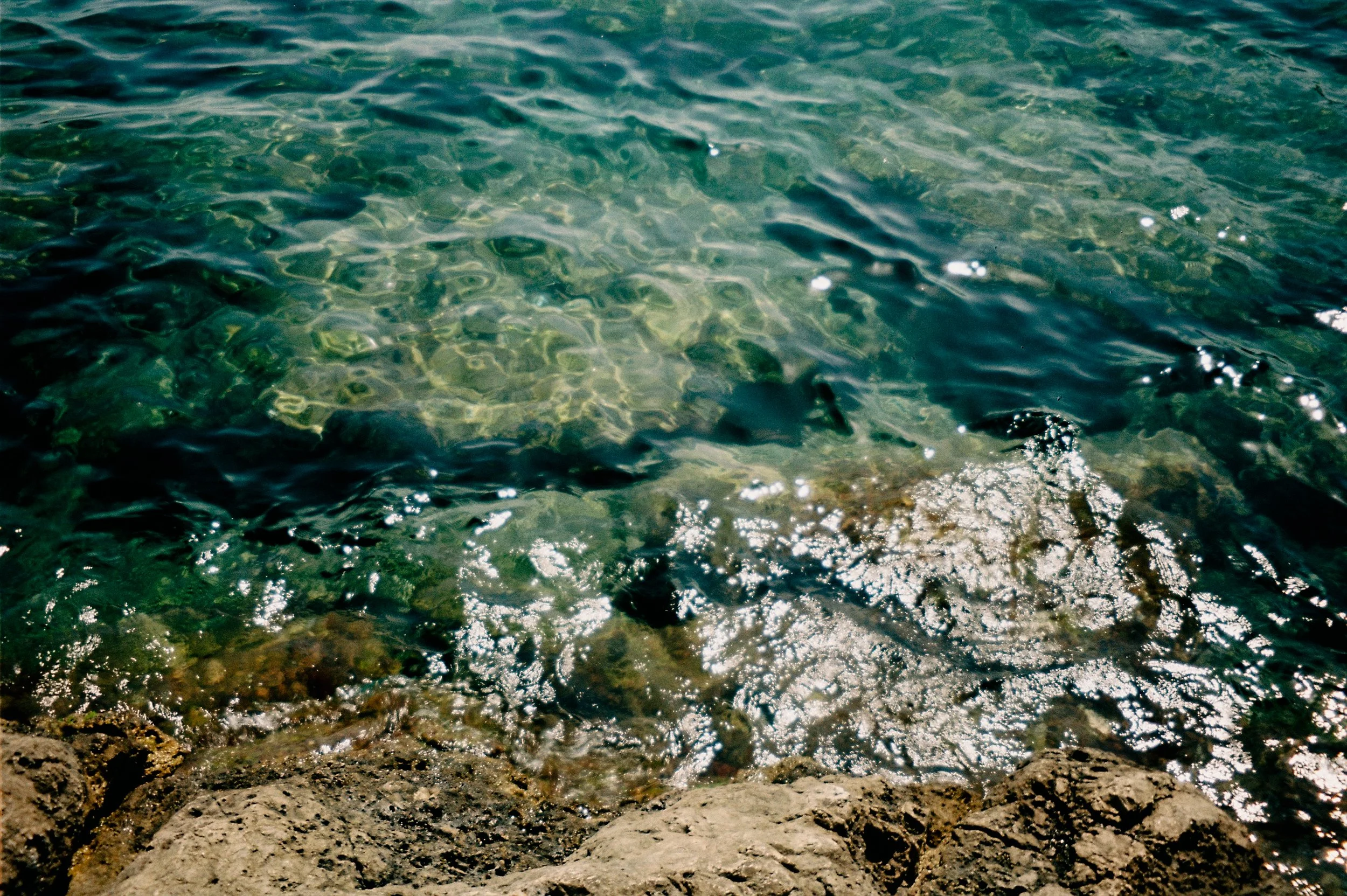 Rocky shoreline with clear, greenish water and sunlight reflections.