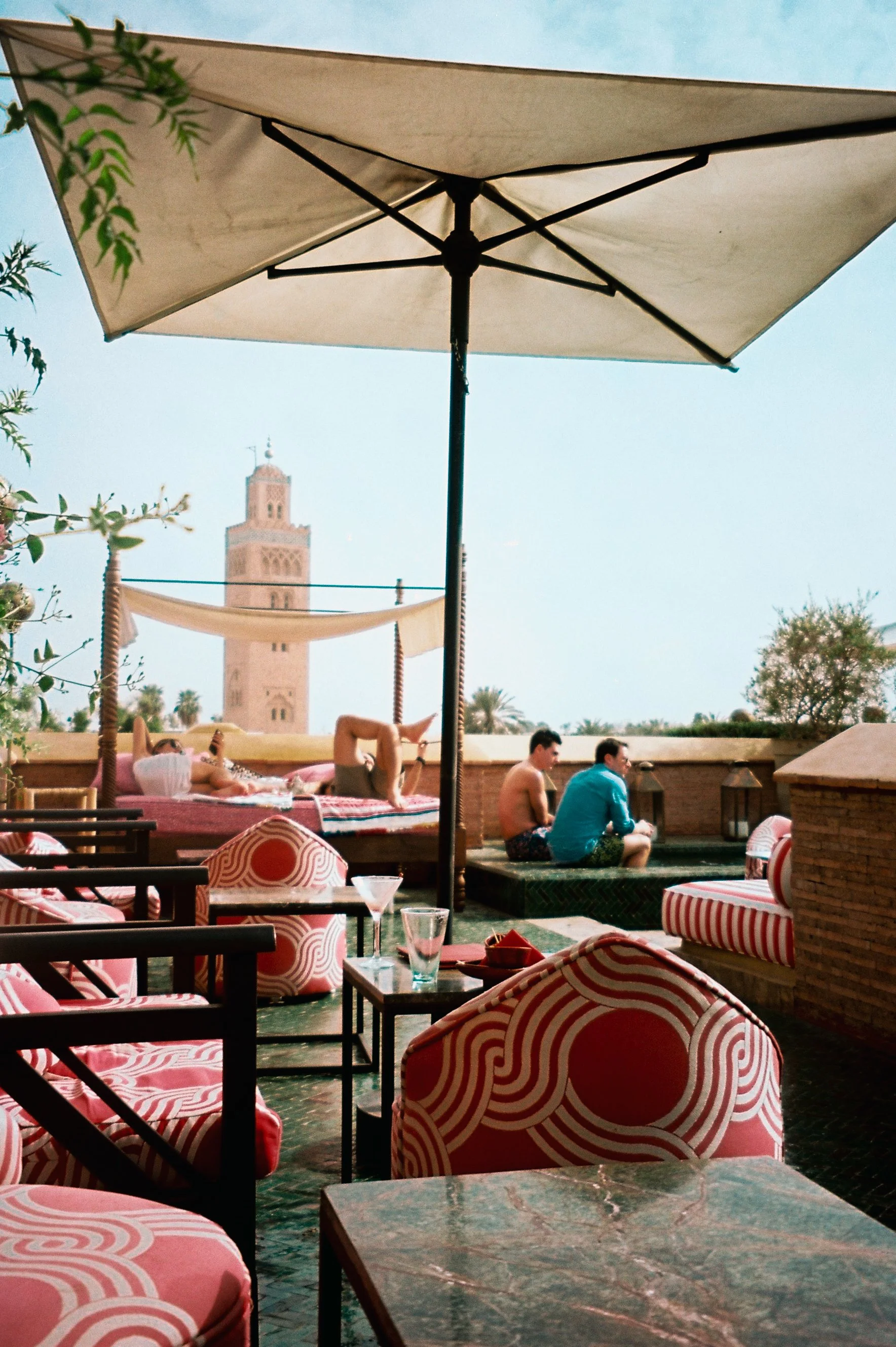 People relaxing on a rooftop terrace with a view of a historic tower in the background, shaded by a large umbrella.