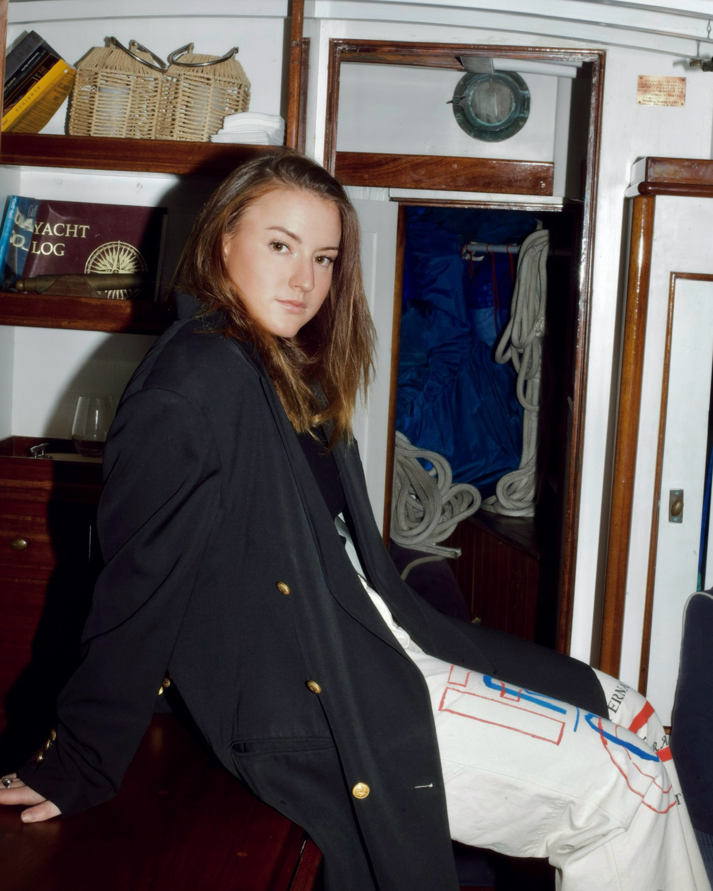 A young woman with brown hair sitting on a wooden surface in a nautical setting, with a nautical logbook, books, and a glass of wine nearby, and nautical ropes and equipment in the background.