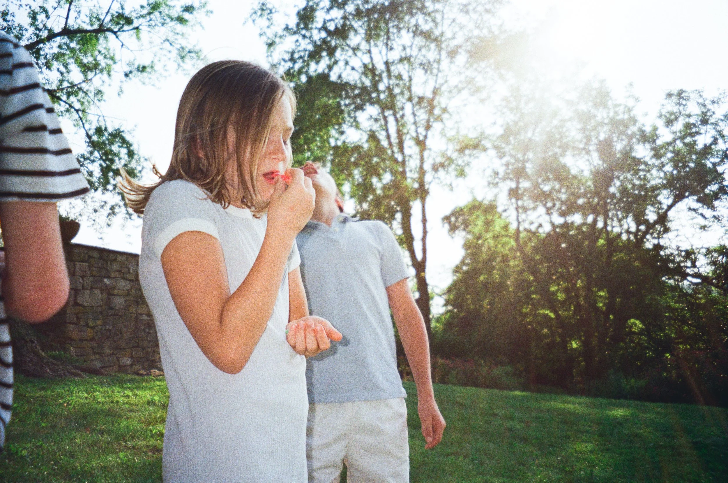 A girl with light brown hair eating a cherry outdoors on a sunny day with trees and grass in the background.