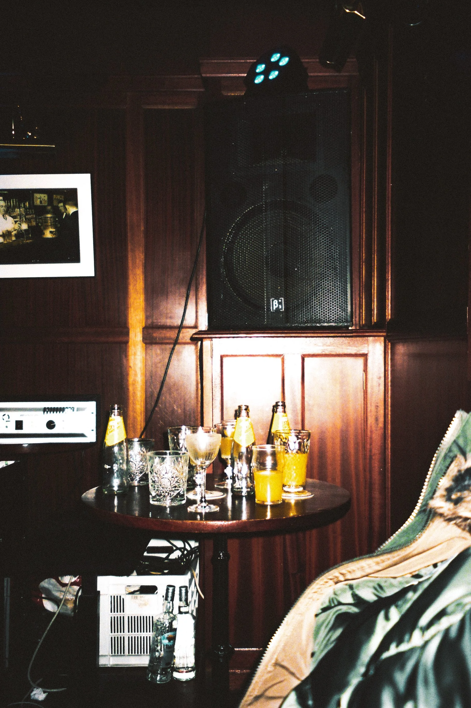 A dark wooden-paneled room with a table holding various glasses and bottles, a large speaker, and a television, in a bar or lounge setting.